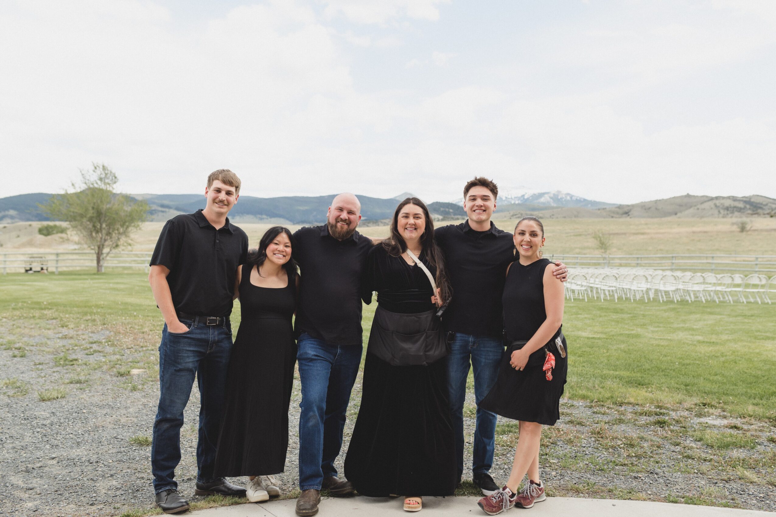 Group of six people standing outdoors on grass with open landscape in background, smiling, dressed casually.