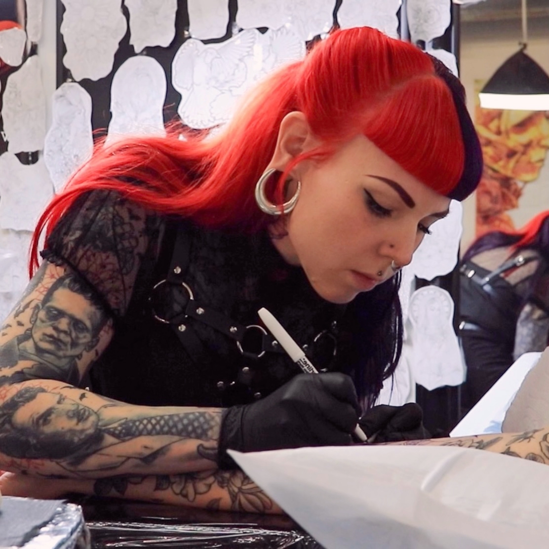Woman with red and black hair, tattoos, and large earrings, writing with a marker on paper, in a tattoo studio.
