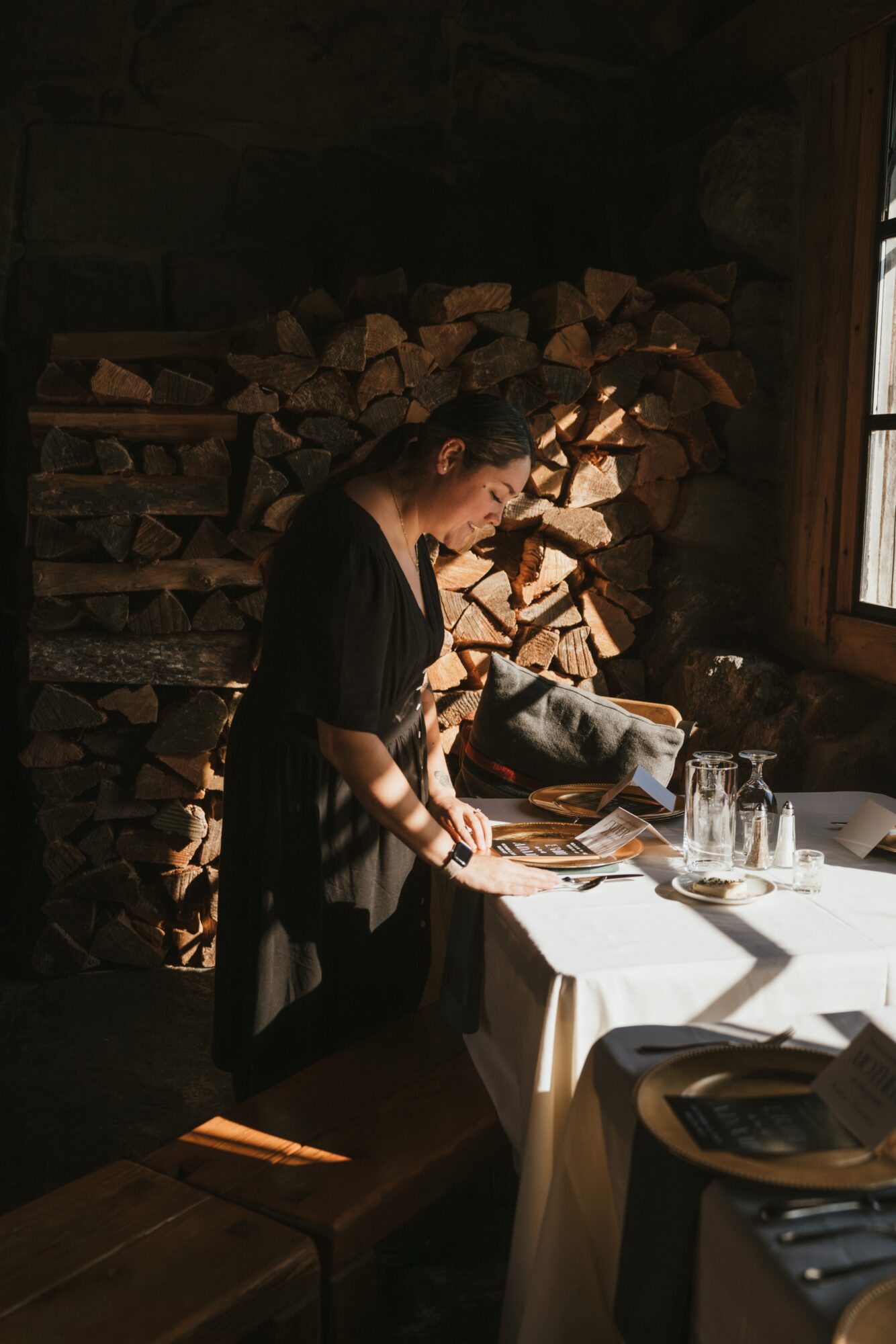 Woman preparing food at a table in a rustic kitchen with stacked firewood and sunlight streaming through a window.