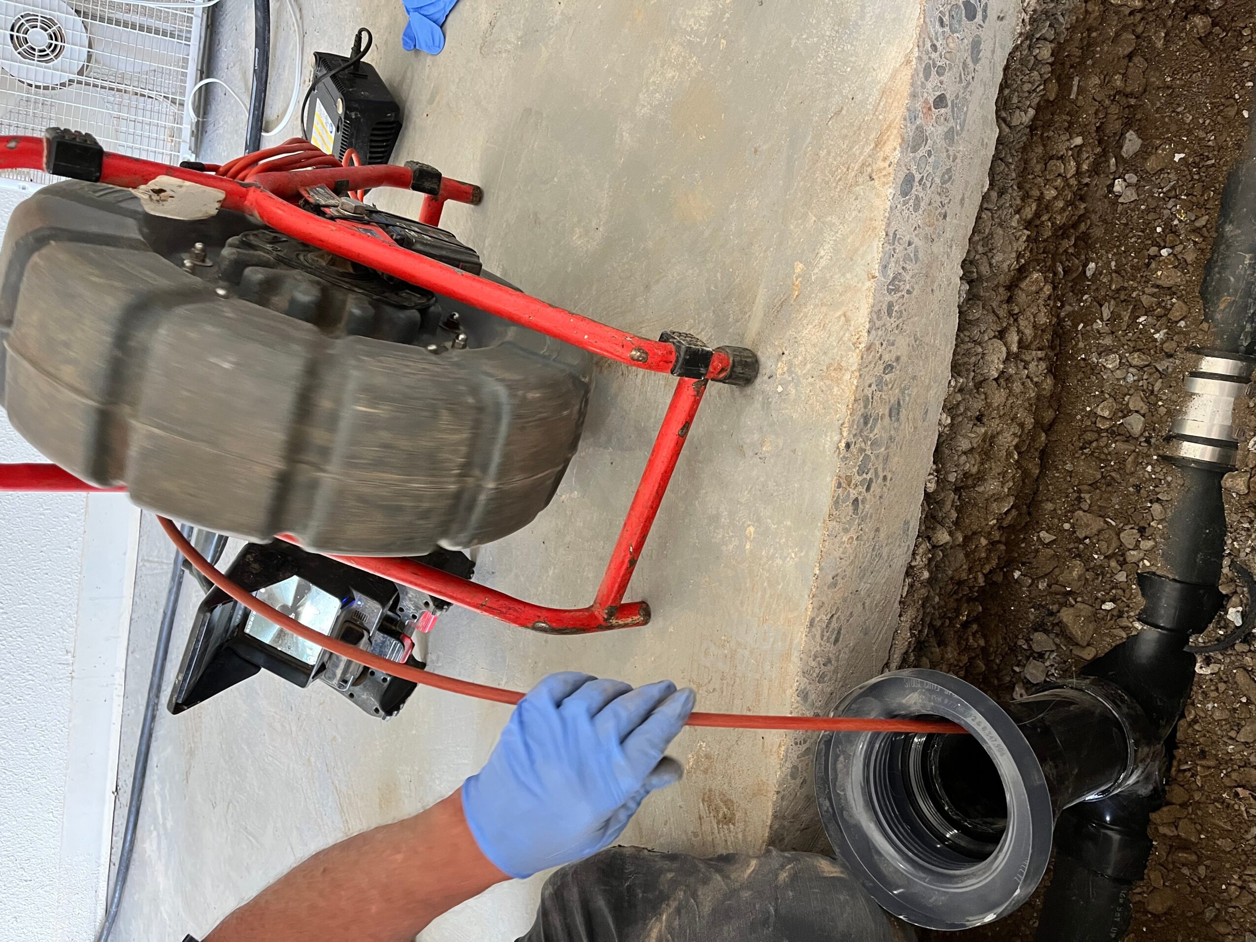 Person wearing blue gloves working on a black pipe with a red tool, concrete surface, and soil nearby.