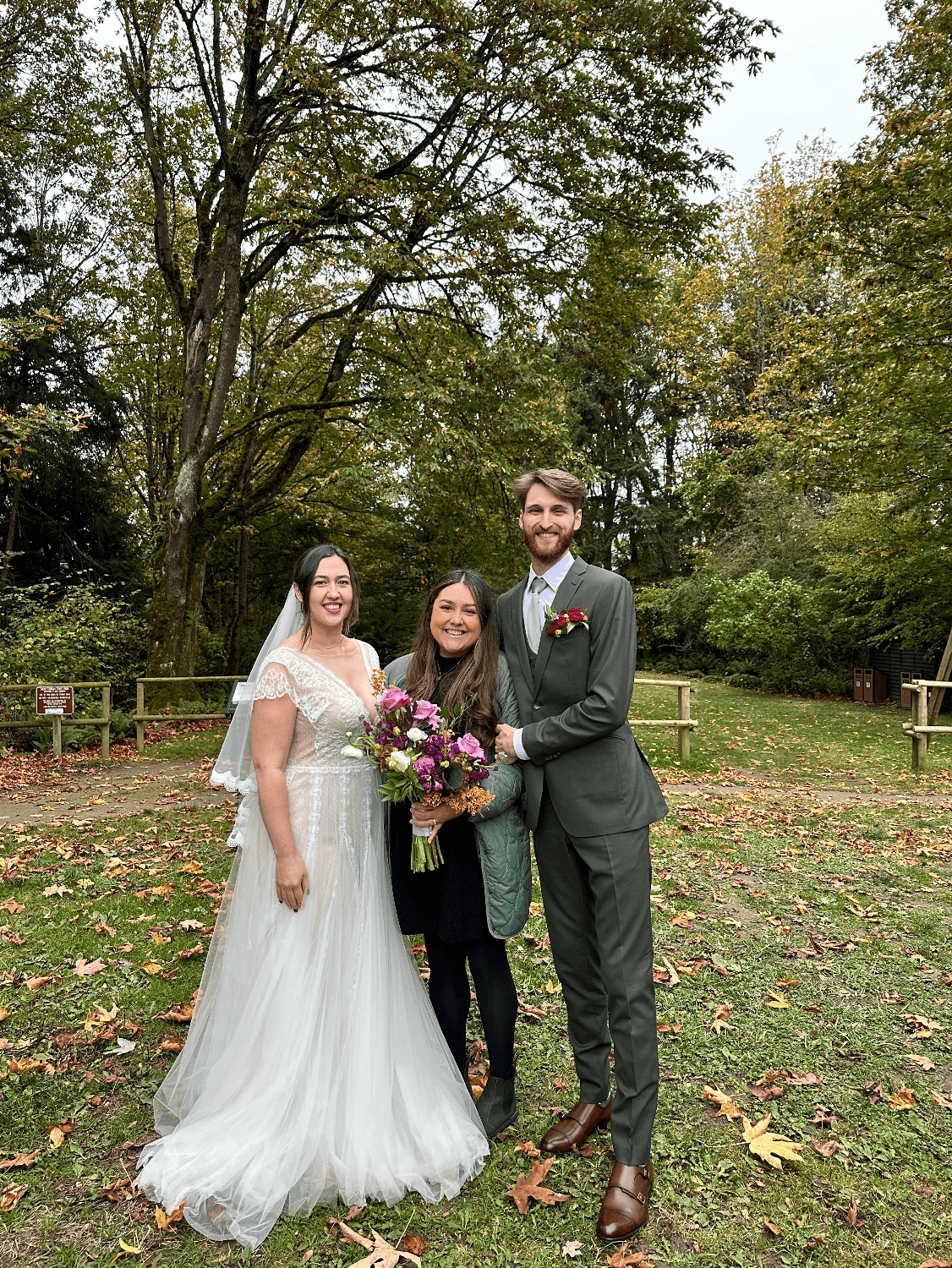 Three people standing outdoors on grass with trees in the background, two women in wedding dress and a man in suit, smiling.