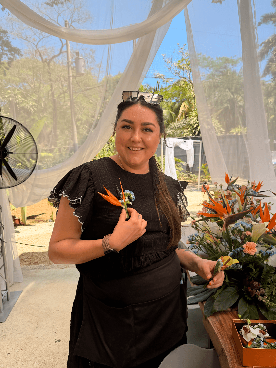 Woman in black dress holding a flower, standing near a table with flowers, outdoors with trees and a white curtain behind.