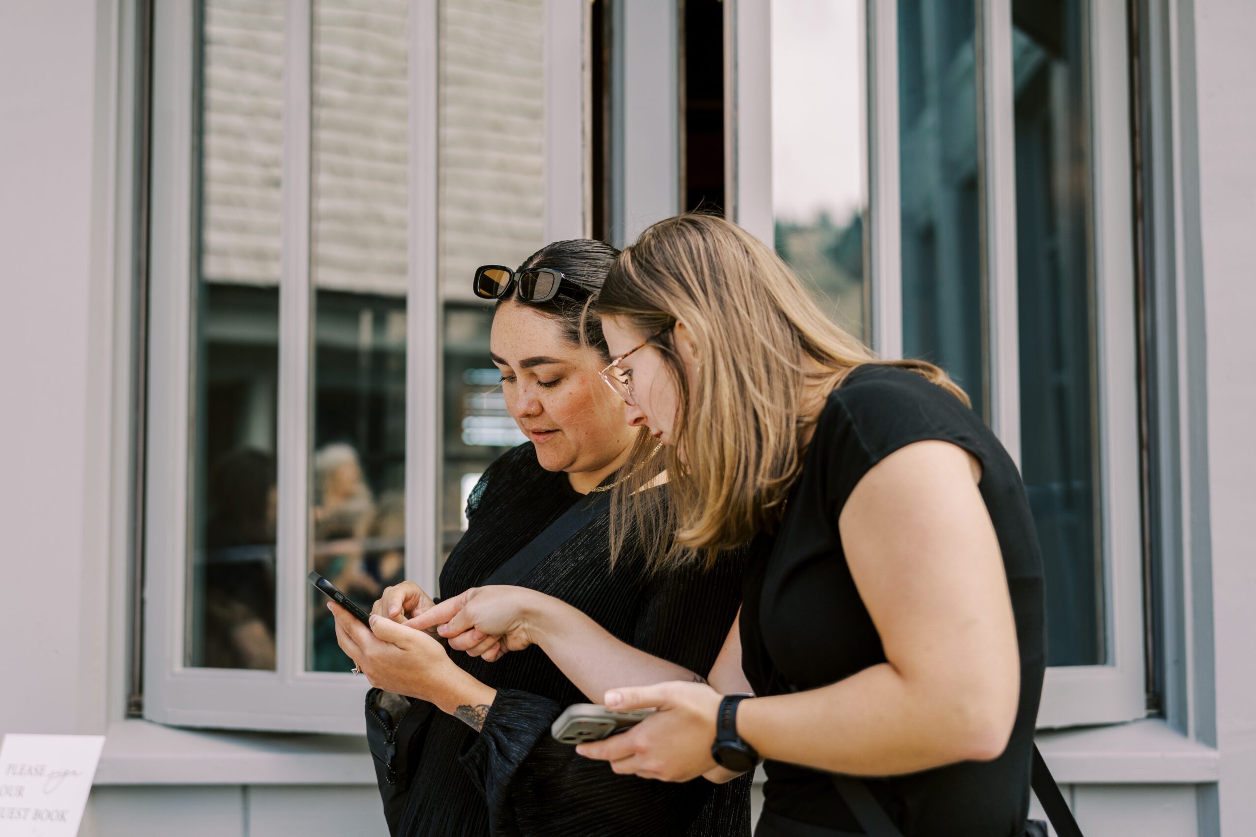 Two women look at a phone together outside a building with large windows, one wearing sunglasses on her head.