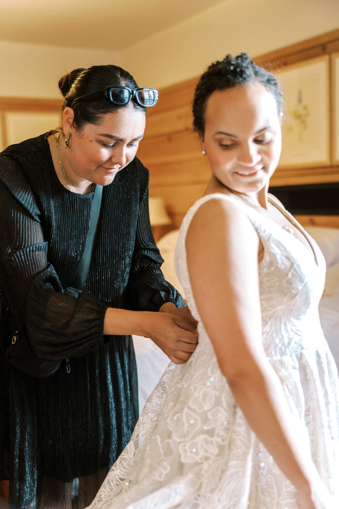 Two women, one in a black dress and sunglasses on her head, helping the other in a white dress, in a wooden-paneled room.