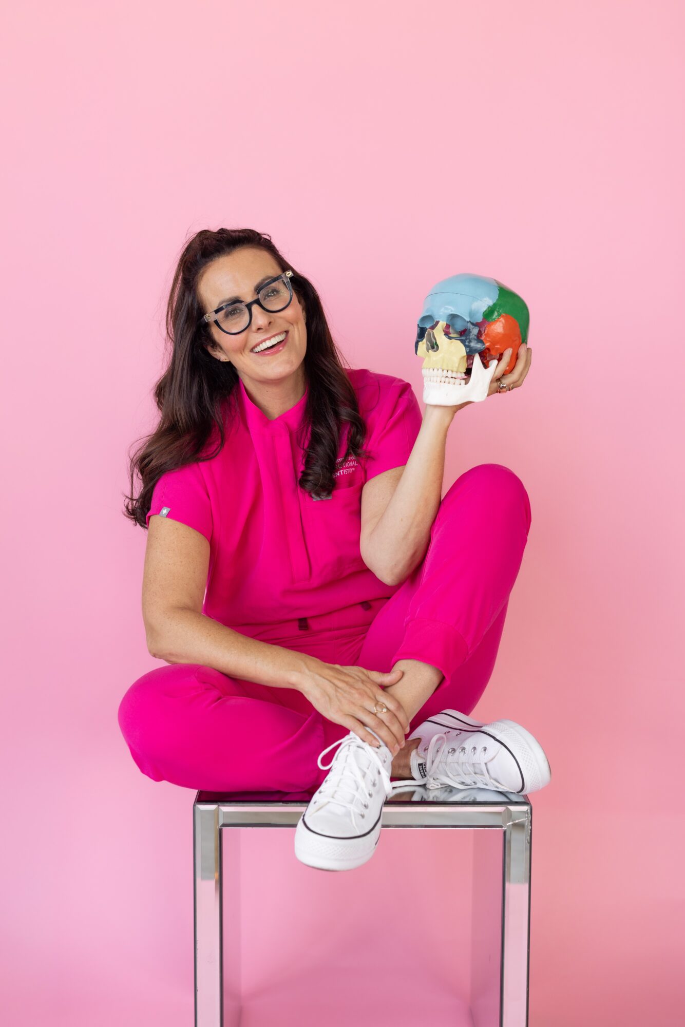 Woman with glasses and long dark hair, wearing pink outfit, sitting on a stool against pink background, holding a colorful globe.