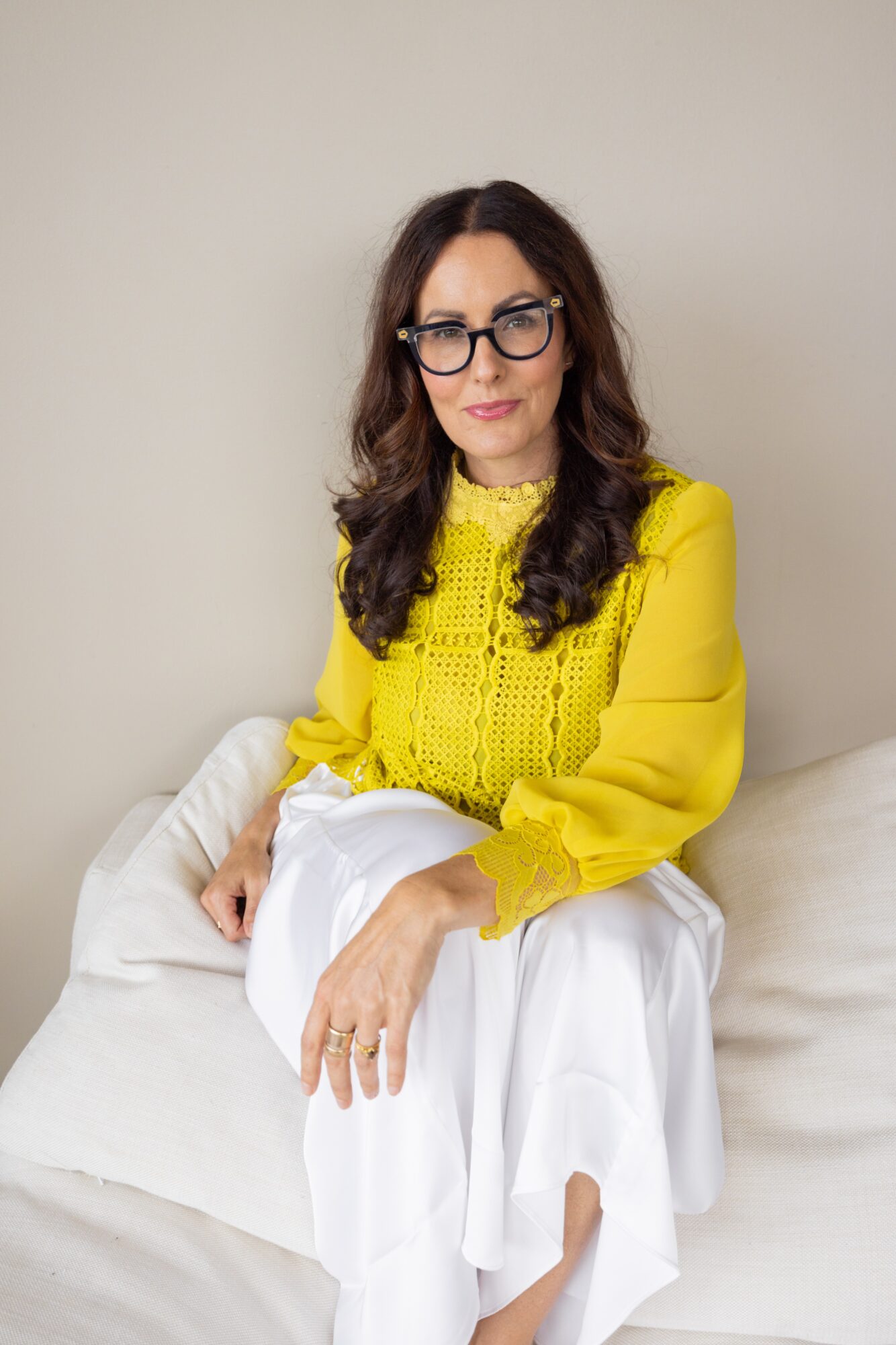 Woman with glasses and dark hair sitting on a white couch, wearing a yellow blouse and white skirt.