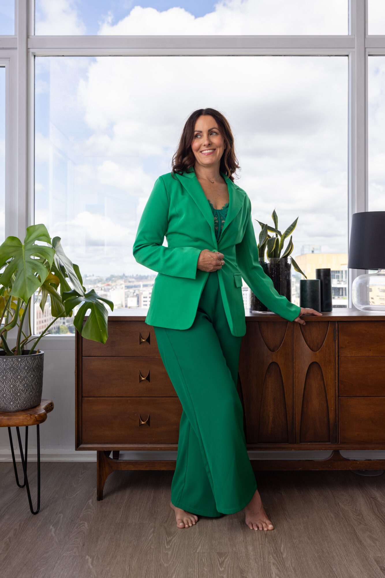 Woman in green suit standing by a desk with plants and a window view of sky and buildings.