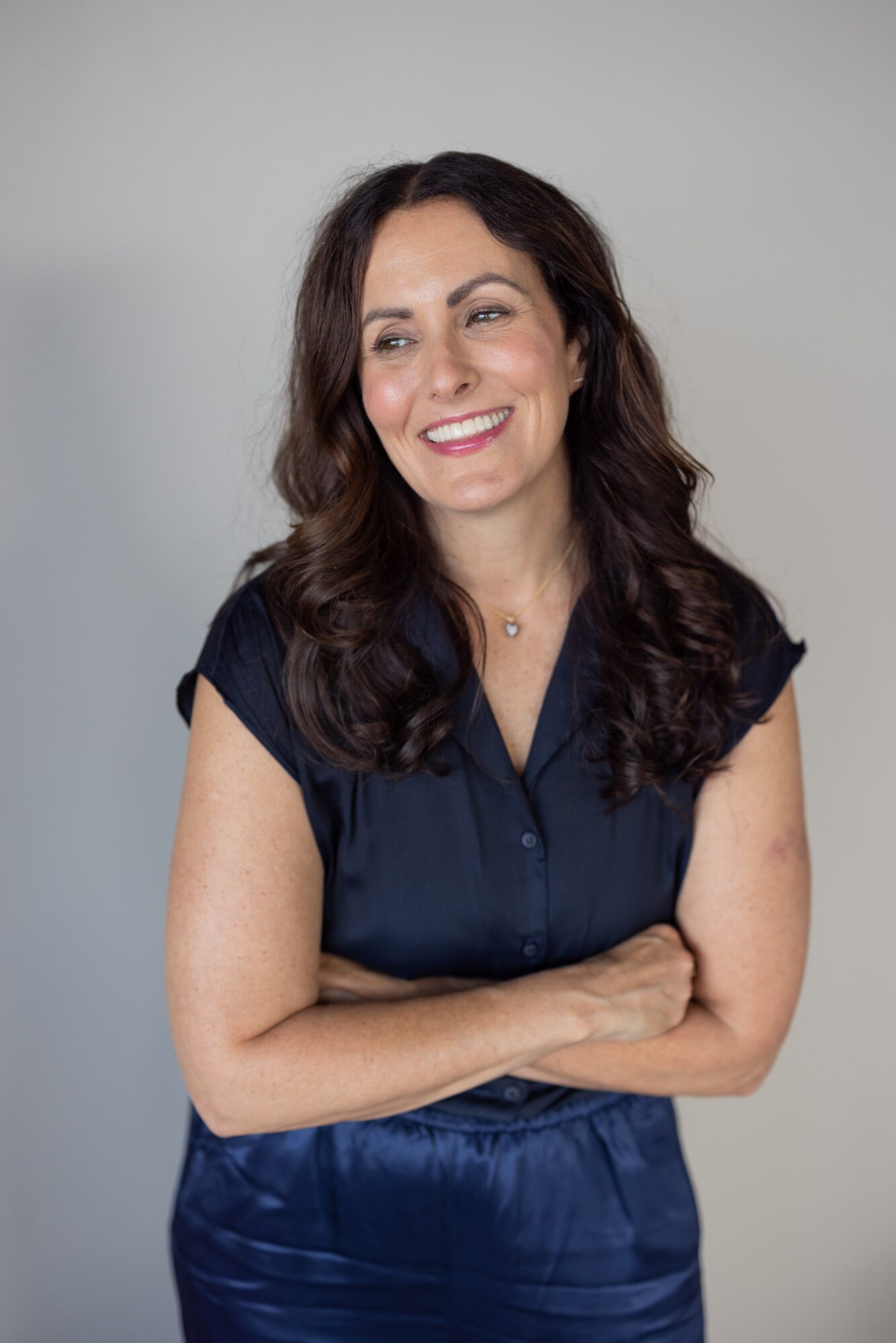 Woman with long dark hair smiling, wearing a navy blue top, arms crossed, against a plain background.