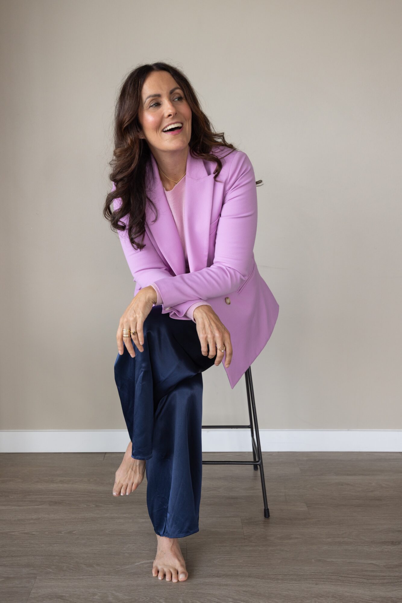 Woman with long dark hair smiling, sitting barefoot on a chair, wearing a pink blazer and dark pants, against a plain background.