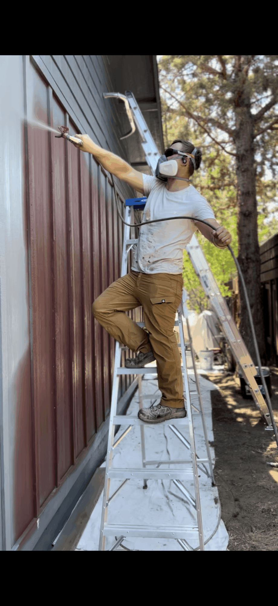 Person standing on a ladder painting a wall outdoors, wearing a mask and cap, with trees in the background.