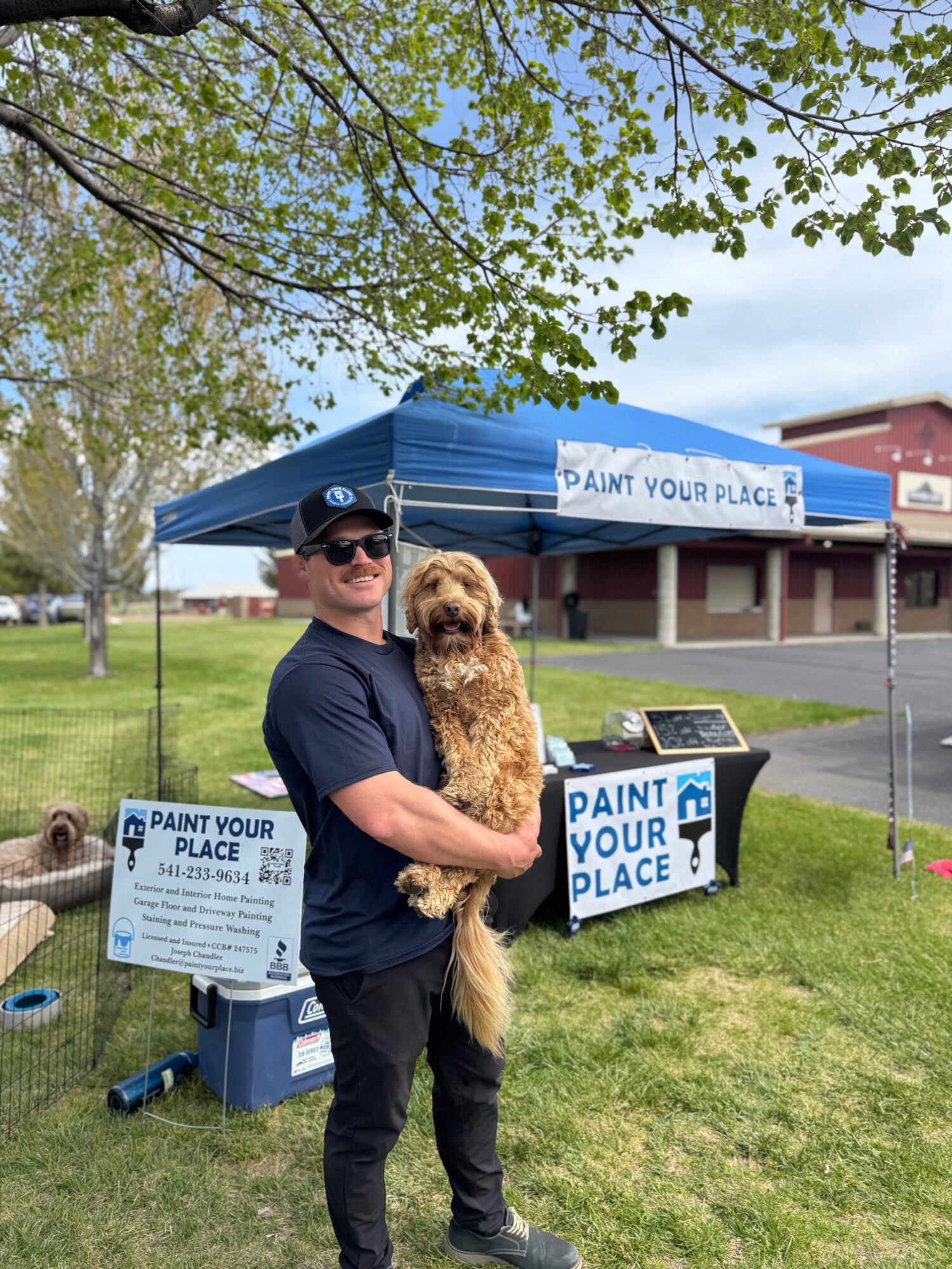 Man holding a dog in front of a tent with signs that say 'Paint Your Place' outdoors on a grassy area.