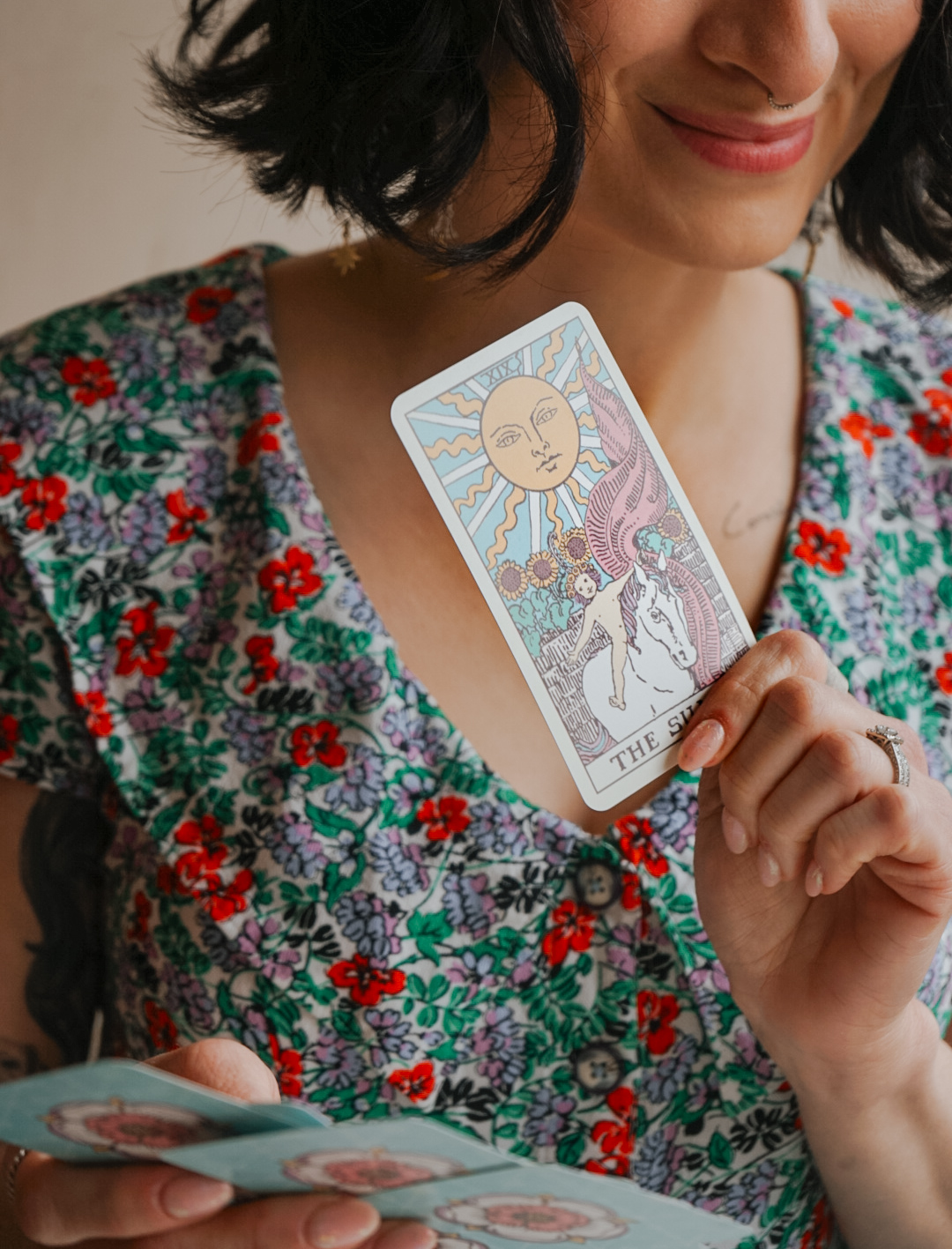 Woman holding a tarot card with a sun and moon illustration, wearing a floral dress, smiling.
