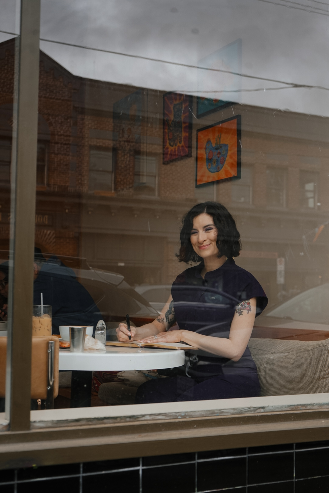 Woman with dark, wavy hair sitting at a table inside a cafe, smiling, with a window reflecting buildings outside.