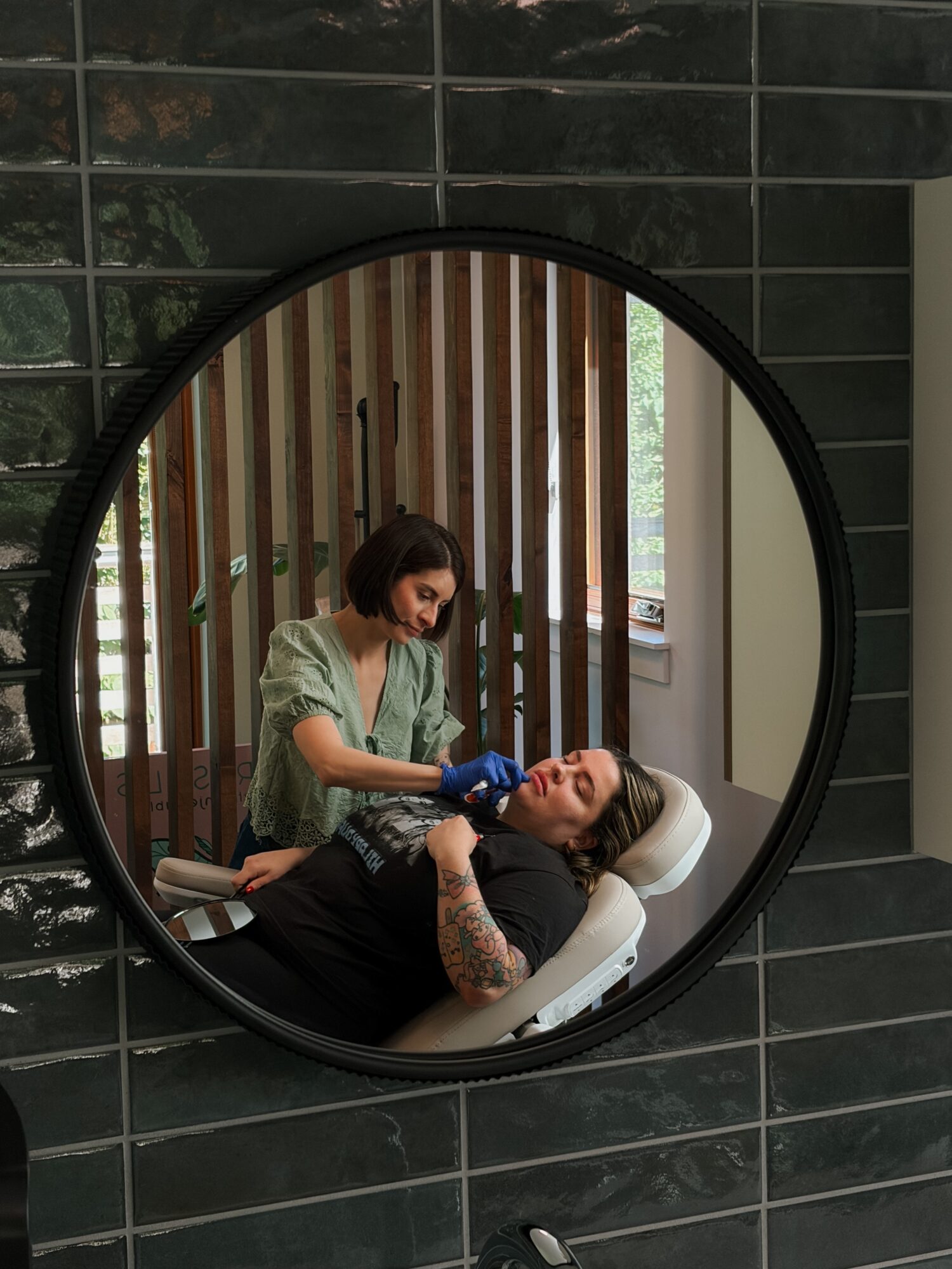 Two women in a salon, one lying on a chair and the other attending to her hair, reflected in a round mirror.