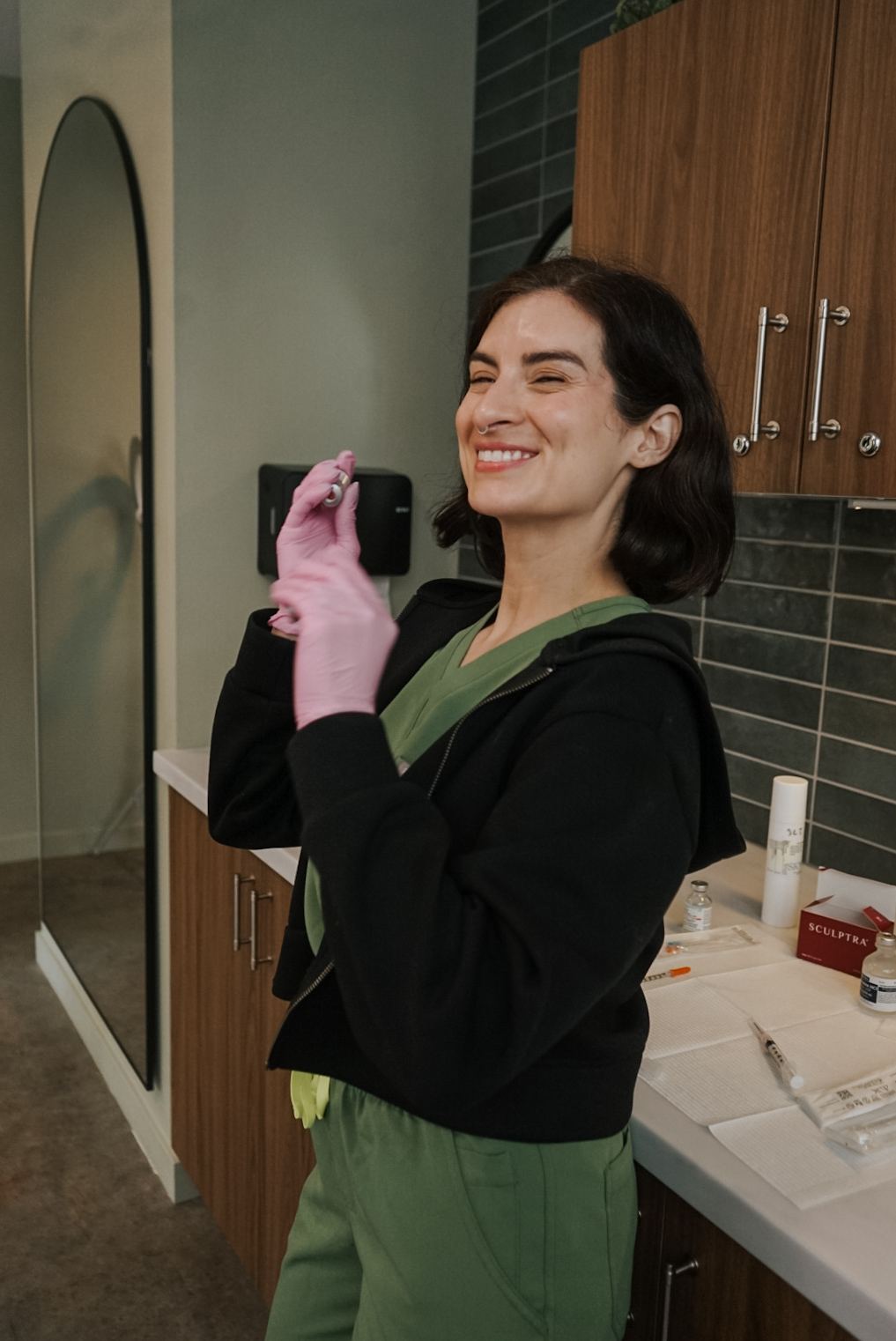 Smiling woman wearing pink gloves in a kitchen with wooden cabinets and a mirror behind her.
