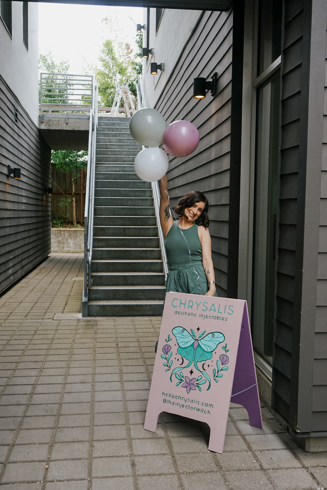 Woman in green dress holding balloons near stairs outside building, with a signboard displaying a butterfly illustration.