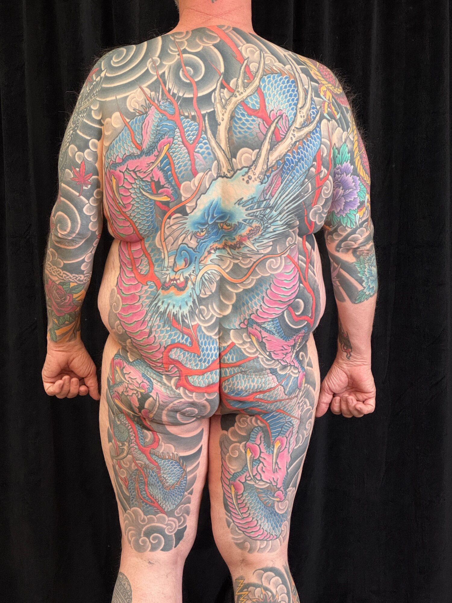Person lying on side with full-body tattoos, including a dragon and clouds, on a black background.