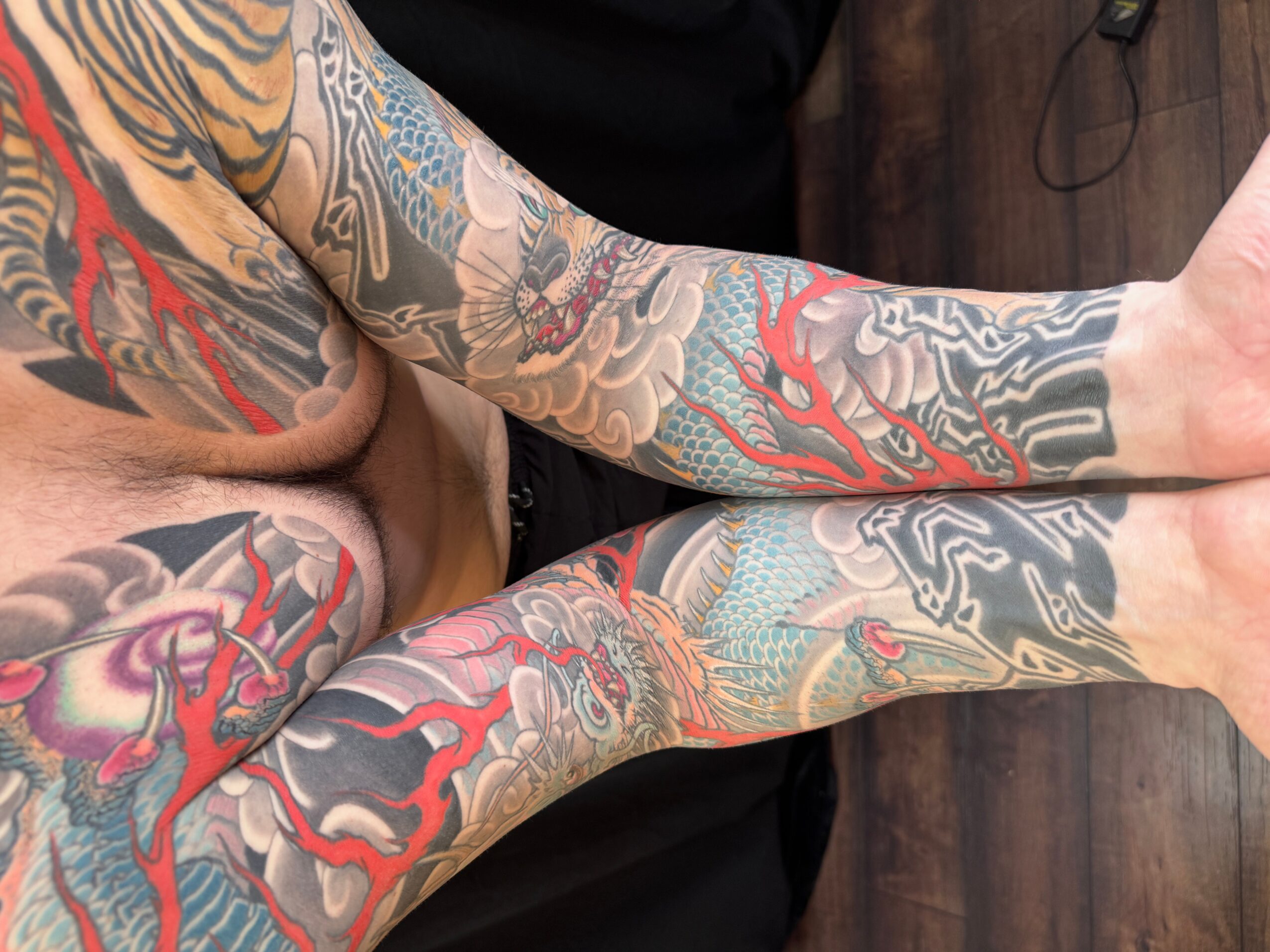 Person with colorful tattoos on both arms, lying down on a dark wooden floor, with arms extended and hands visible.