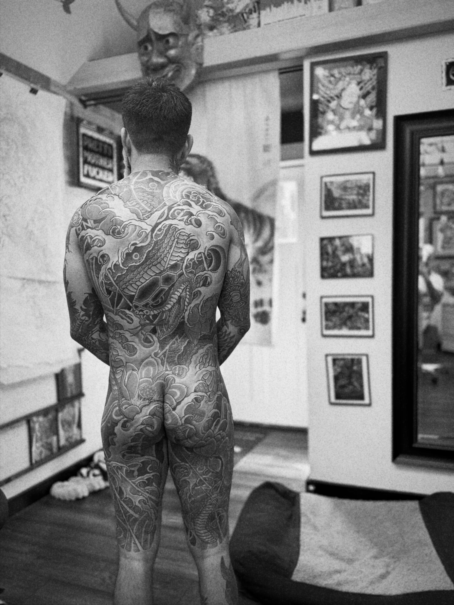 Back view of a person with intricate tattoos standing in a room with framed pictures on the wall.