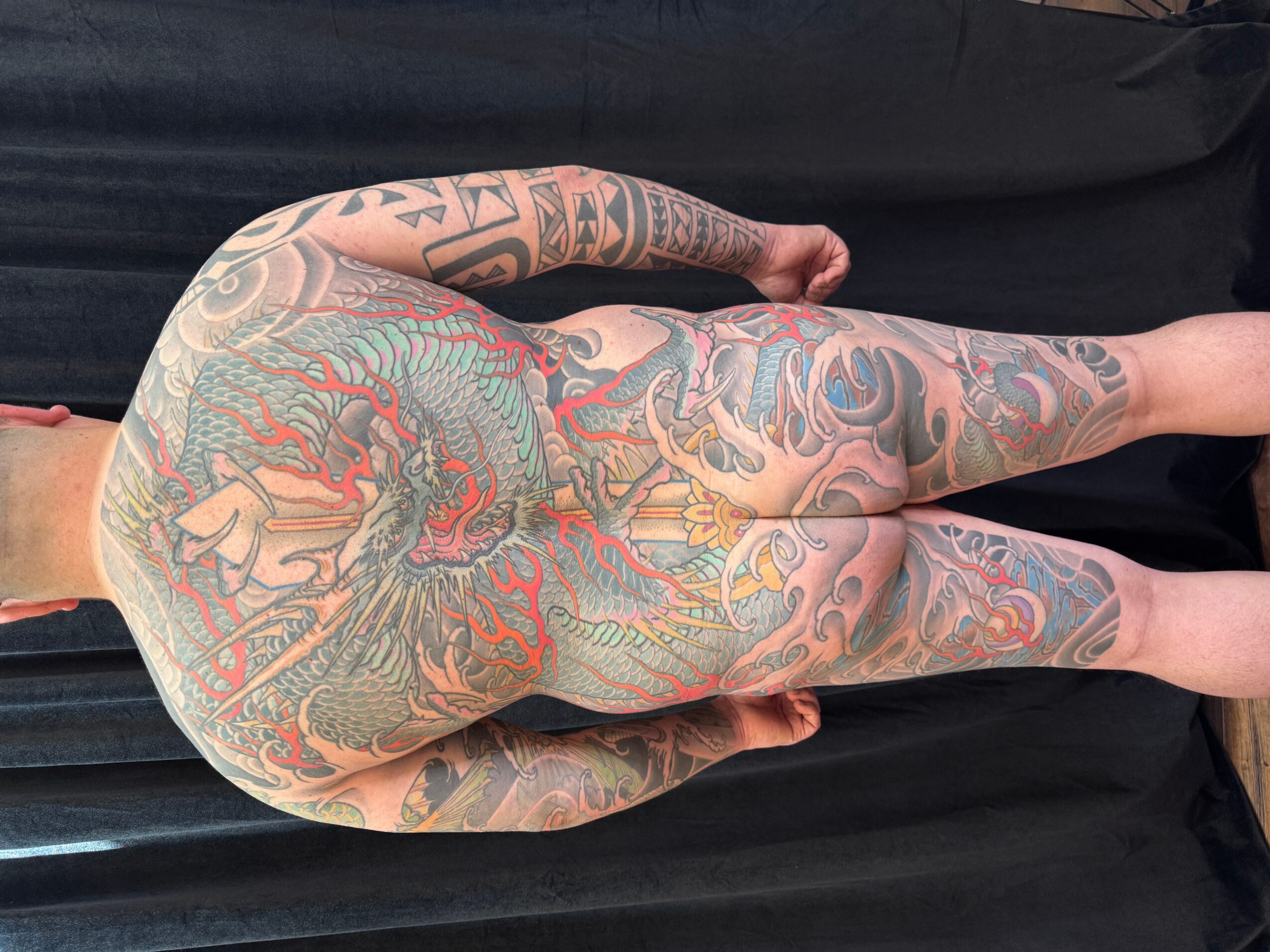 Person with extensive colorful tattoos covering back, arms, and legs, lying face down on black surface.