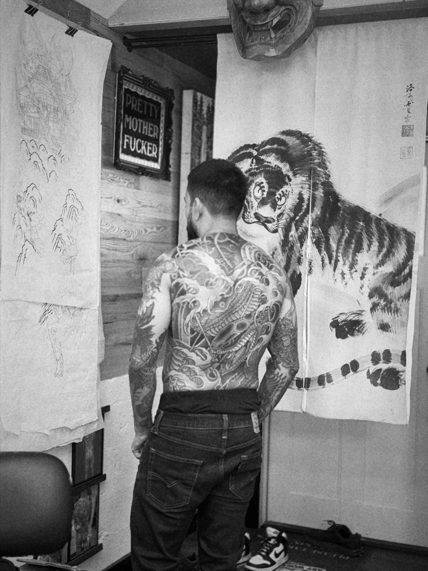 Person with tattoos on back standing in front of tiger painting on wall, facing away from camera, in a room with various artworks.