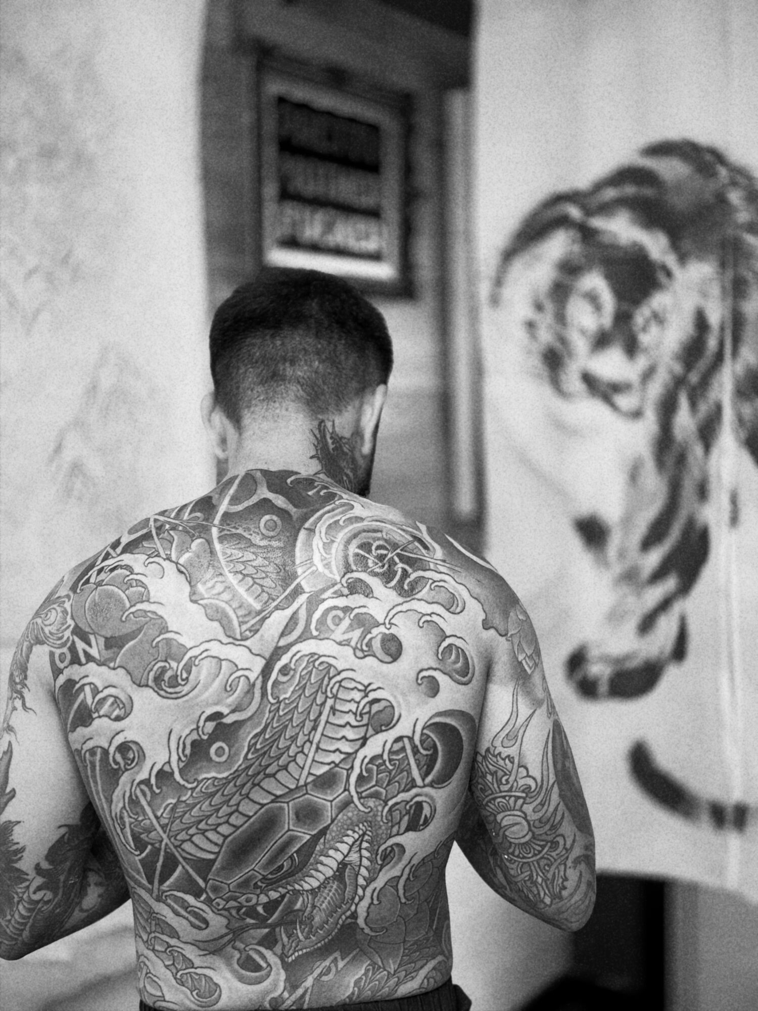 Back of a shirtless man with detailed tattoos on his back and arms, facing a tiger painting on the wall.