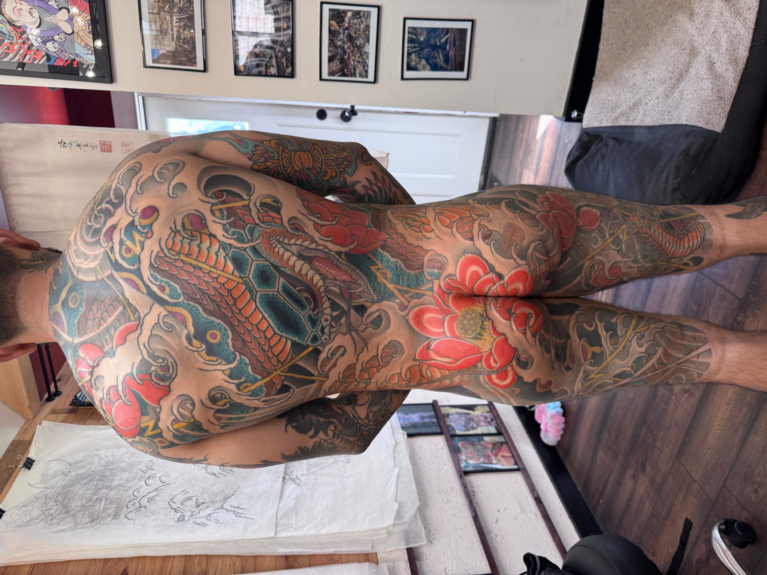 Person with extensive colorful tattoos on back, arms, and legs, lying on side in a room with framed pictures on wall.