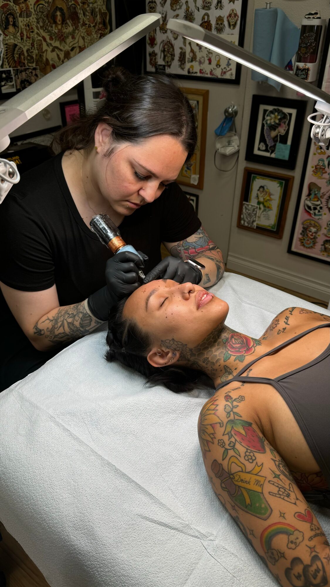 Tattooed woman lying on a bed receiving a cosmetic procedure from a professional with tattoos, in a decorated room.