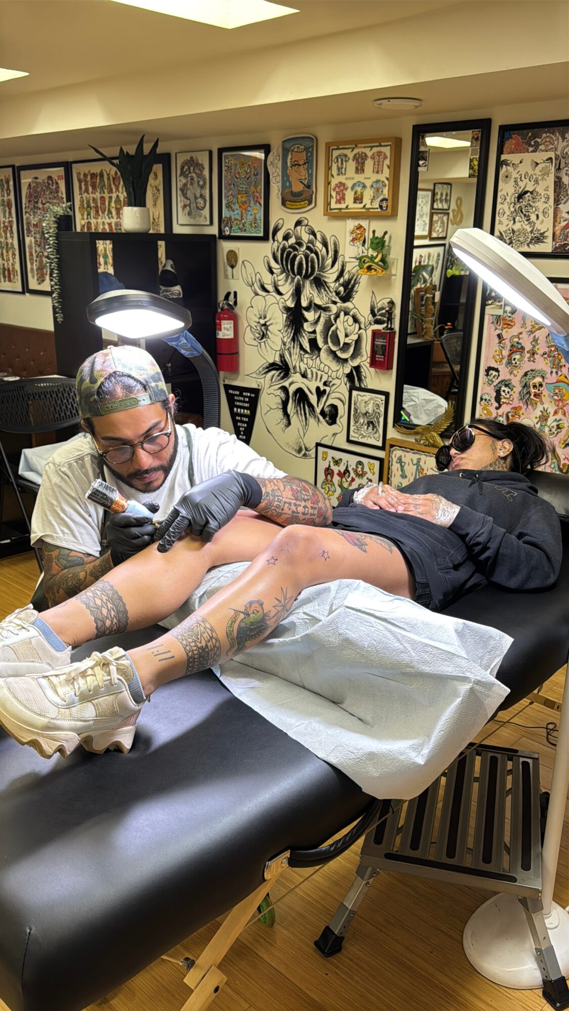 Tattoo artist working on client's leg in tattoo studio with framed artwork on walls.
