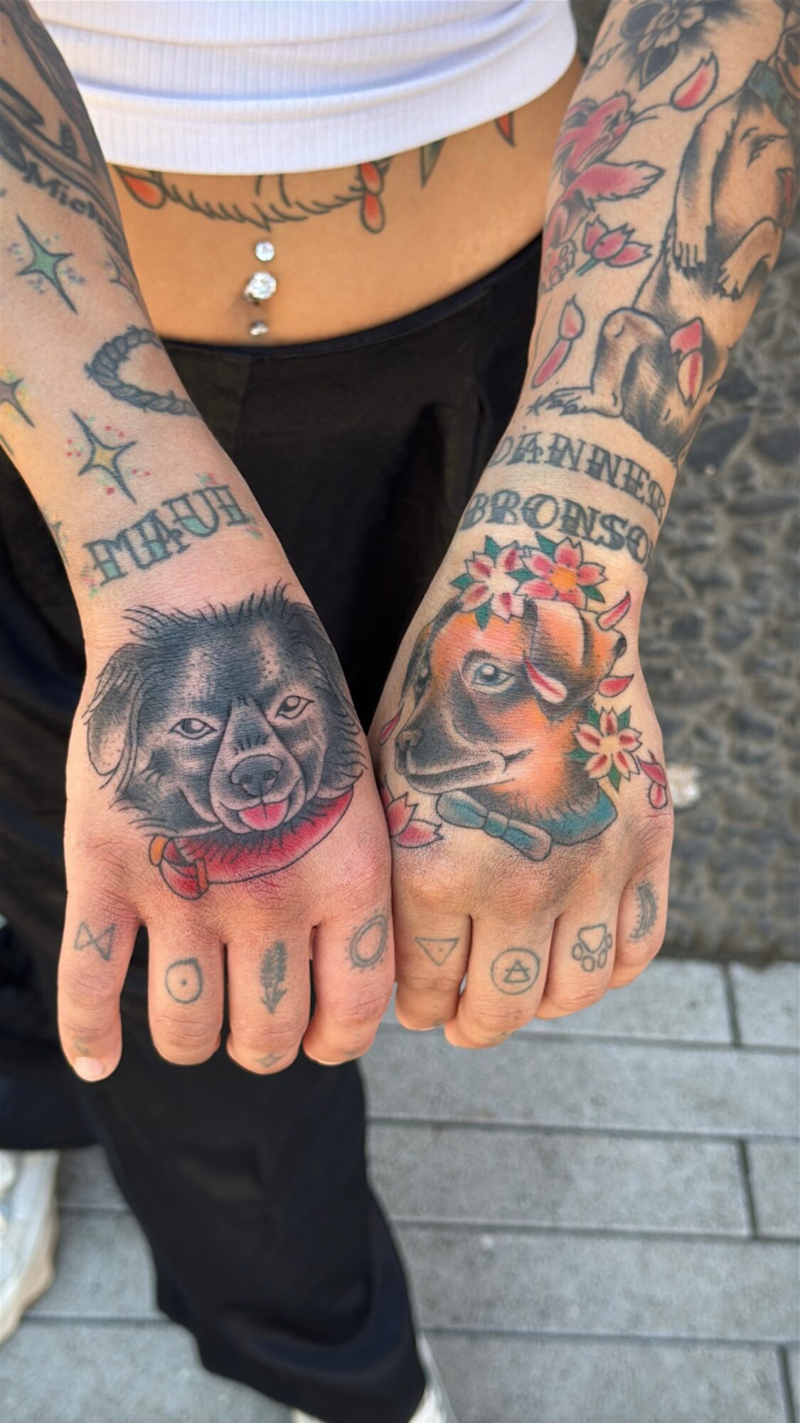 Person with colorful tattoos on hands and arms, including animals, text, and decorative elements, wearing a white top and black pants.
