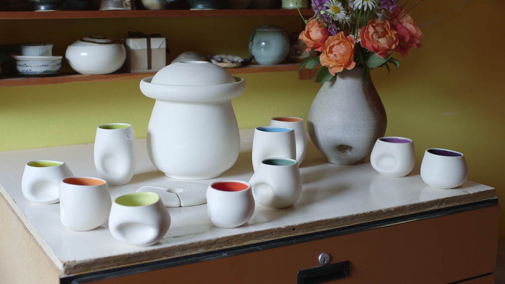 Ceramic cups and vases arranged on a marble surface with a flower arrangement in a vase in the background.