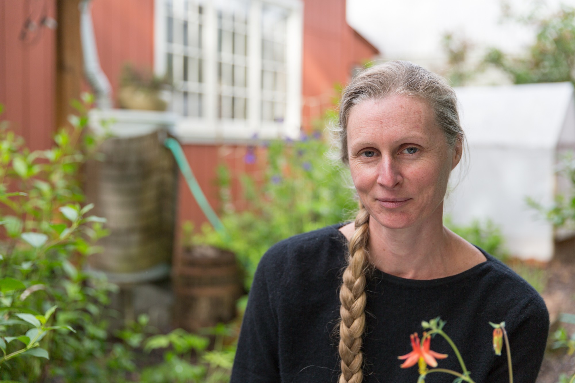 Woman with long braided hair standing outdoors in a garden, looking at the camera, with plants and a house in the background.
