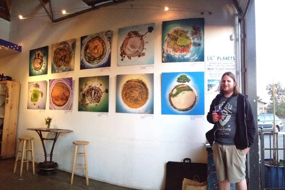 Gallery wall with colorful artwork, a person holding a drink standing nearby, and a small table with stools inside a room.