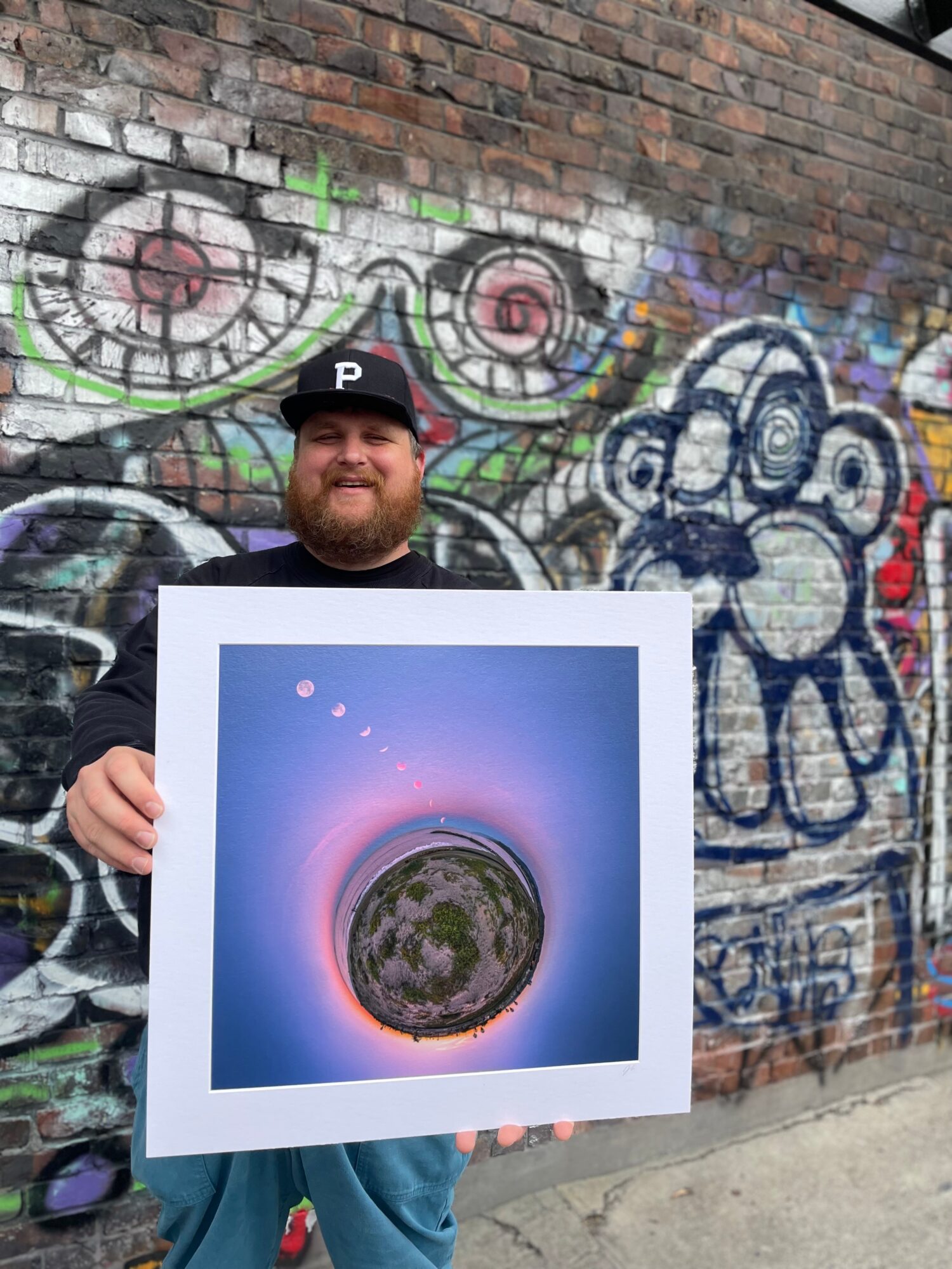 Man with a beard holding a framed artwork in front of a graffiti-covered brick wall.