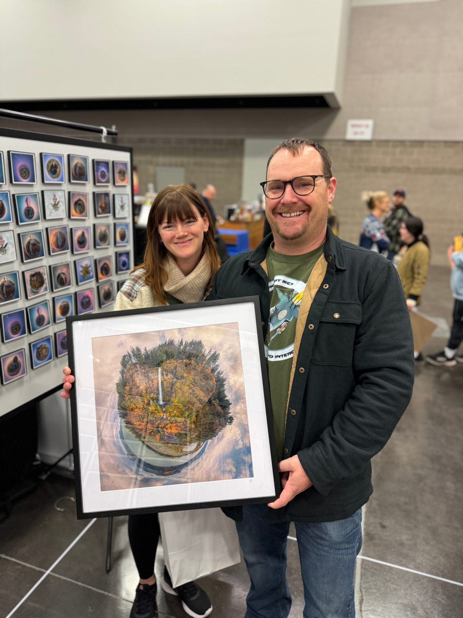 Man and girl holding framed artwork in a gallery, smiling, with other people in background.