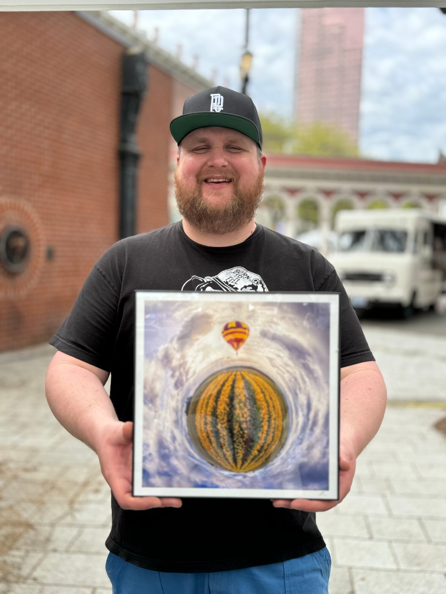 Smiling man with a beard holding a framed artwork outdoors, brick building and trees in background.