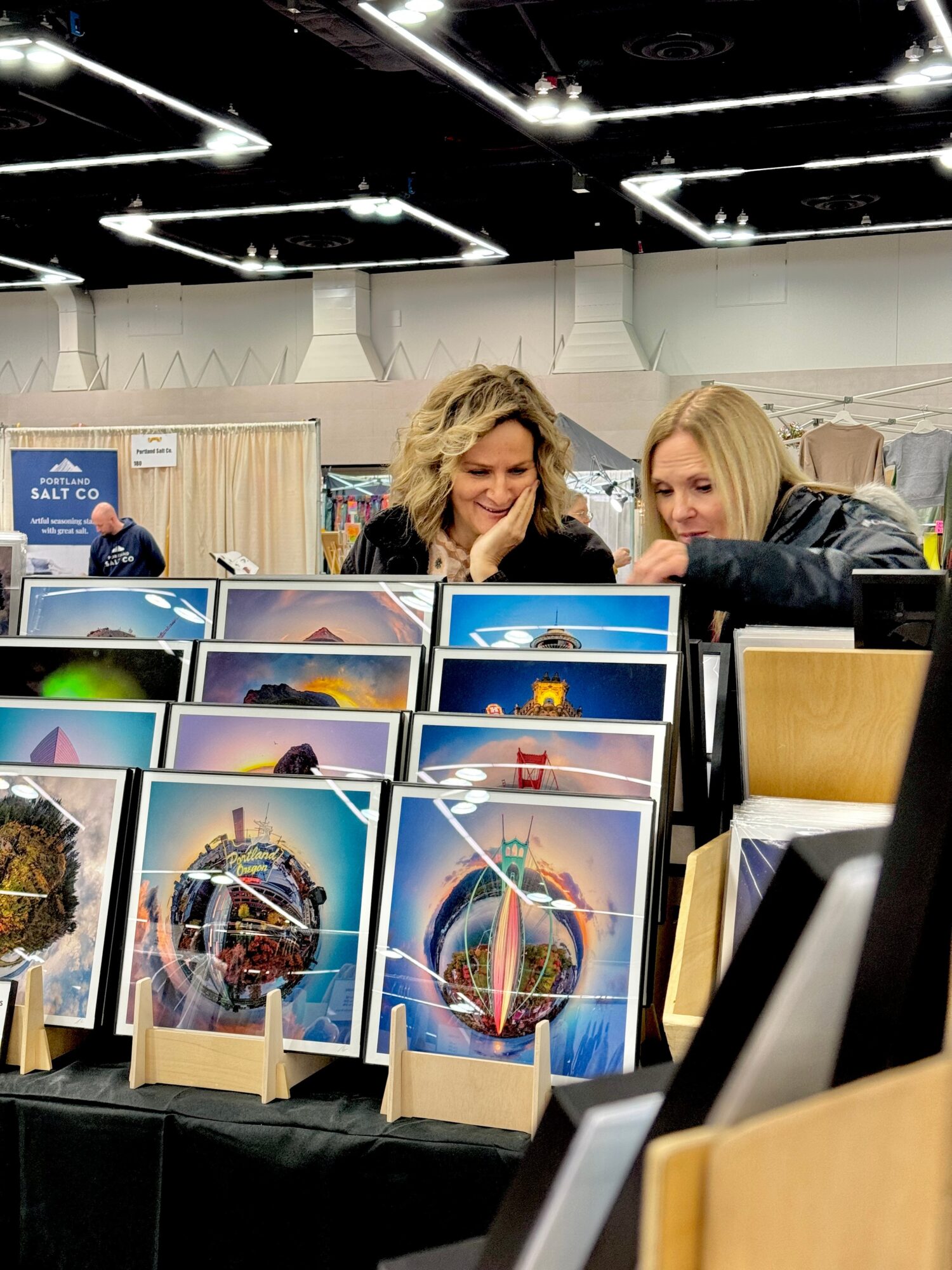 Two women look at colorful artwork displayed on a table at an indoor event, with booths and ceiling lights in background.