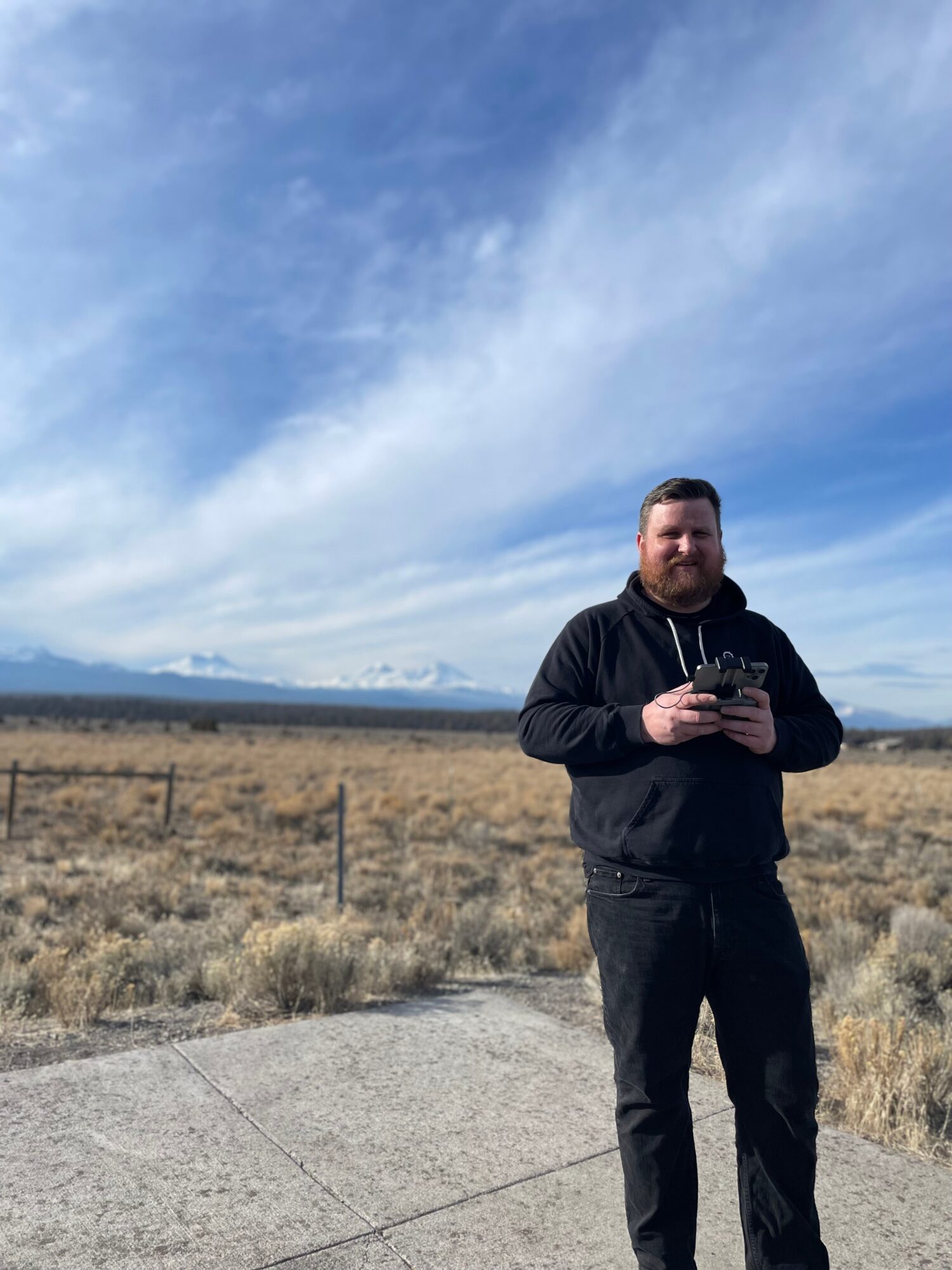 Man standing outdoors in a field, holding a phone, with a blue sky and distant mountains in the background.