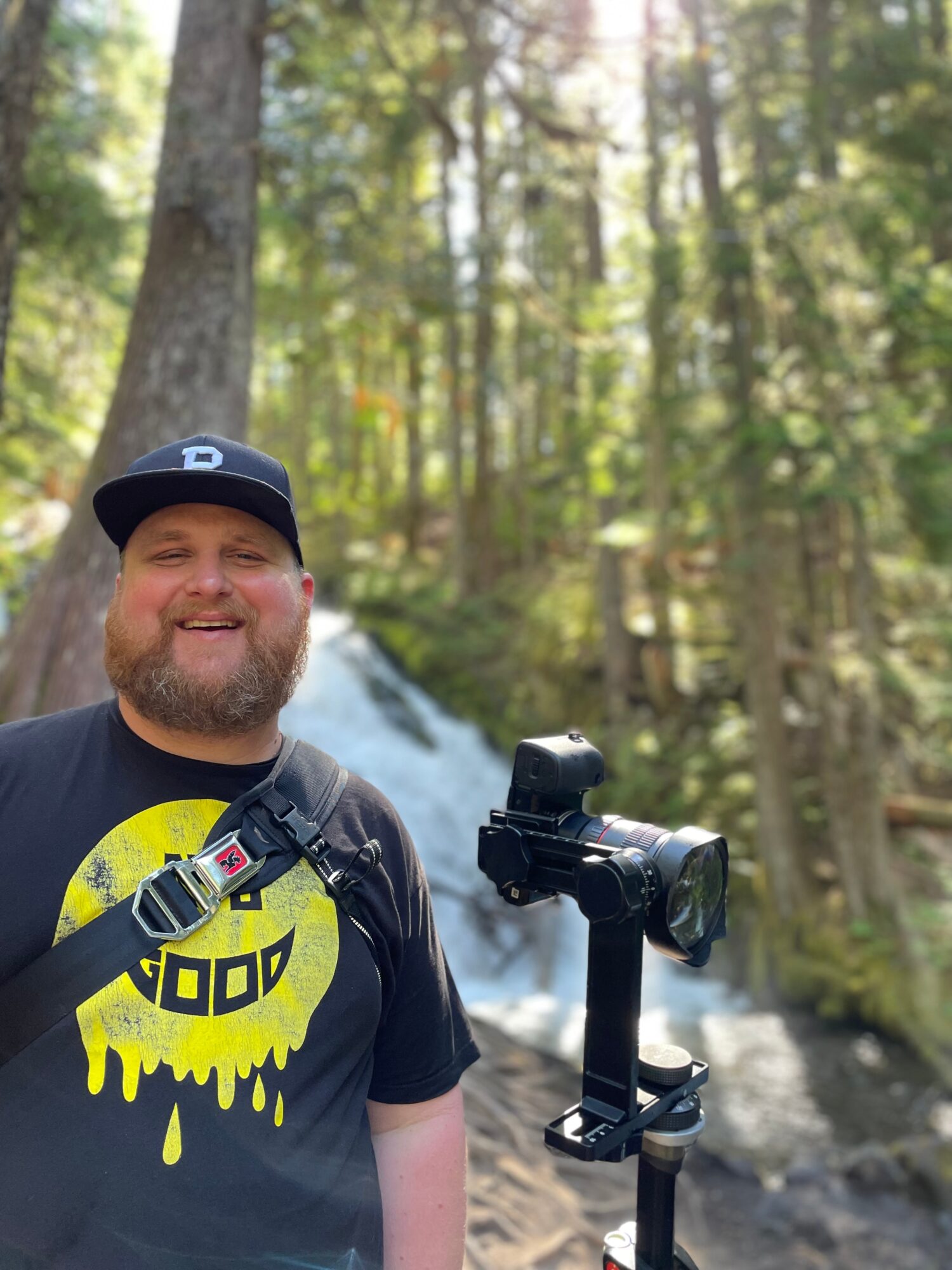 Man with a beard and black cap smiling outdoors near a waterfall in a forest.