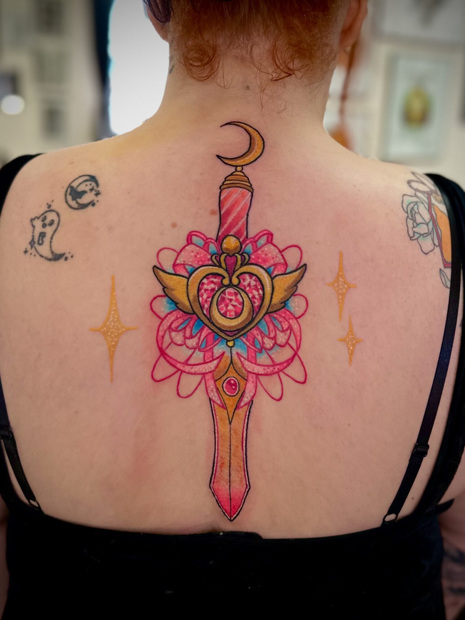 Back of person with colorful dagger tattoo, crescent moon, and star tattoos, with blurred background.