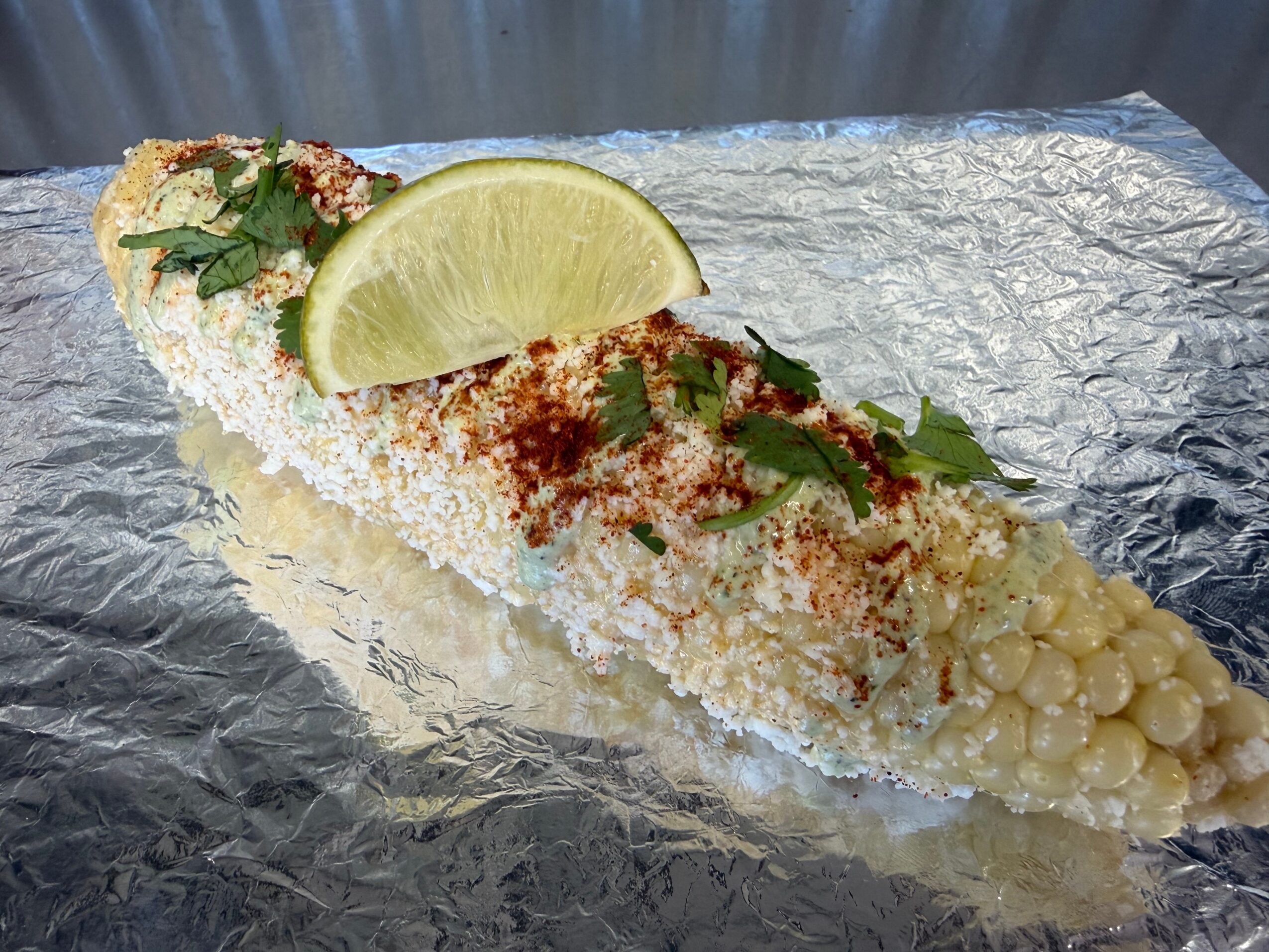 Grilled corn on the cob with lime wedge, cilantro, and spices on aluminum foil.