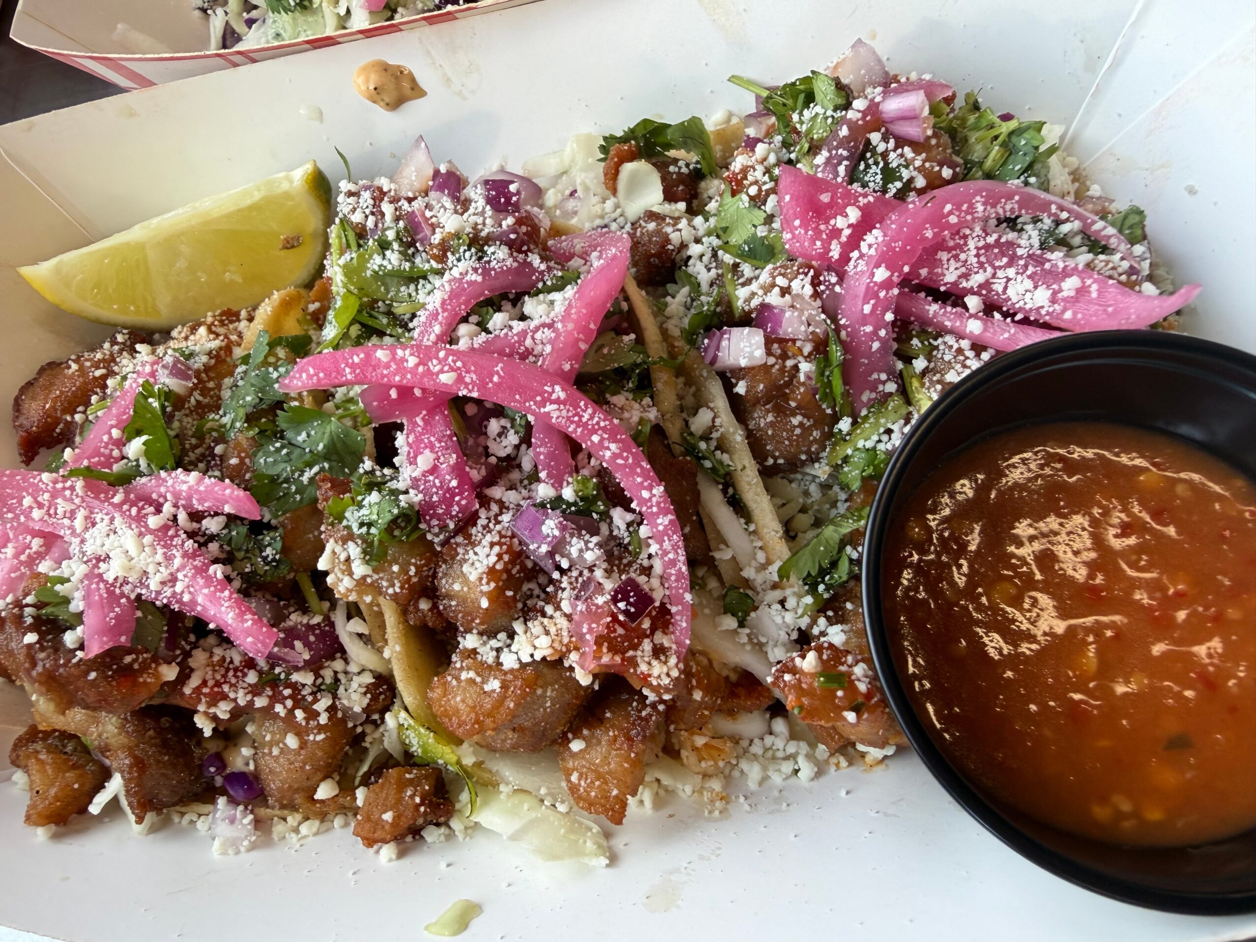 Plate of tacos with pink pickled onions, lime wedge, and a side of red sauce in a black bowl.