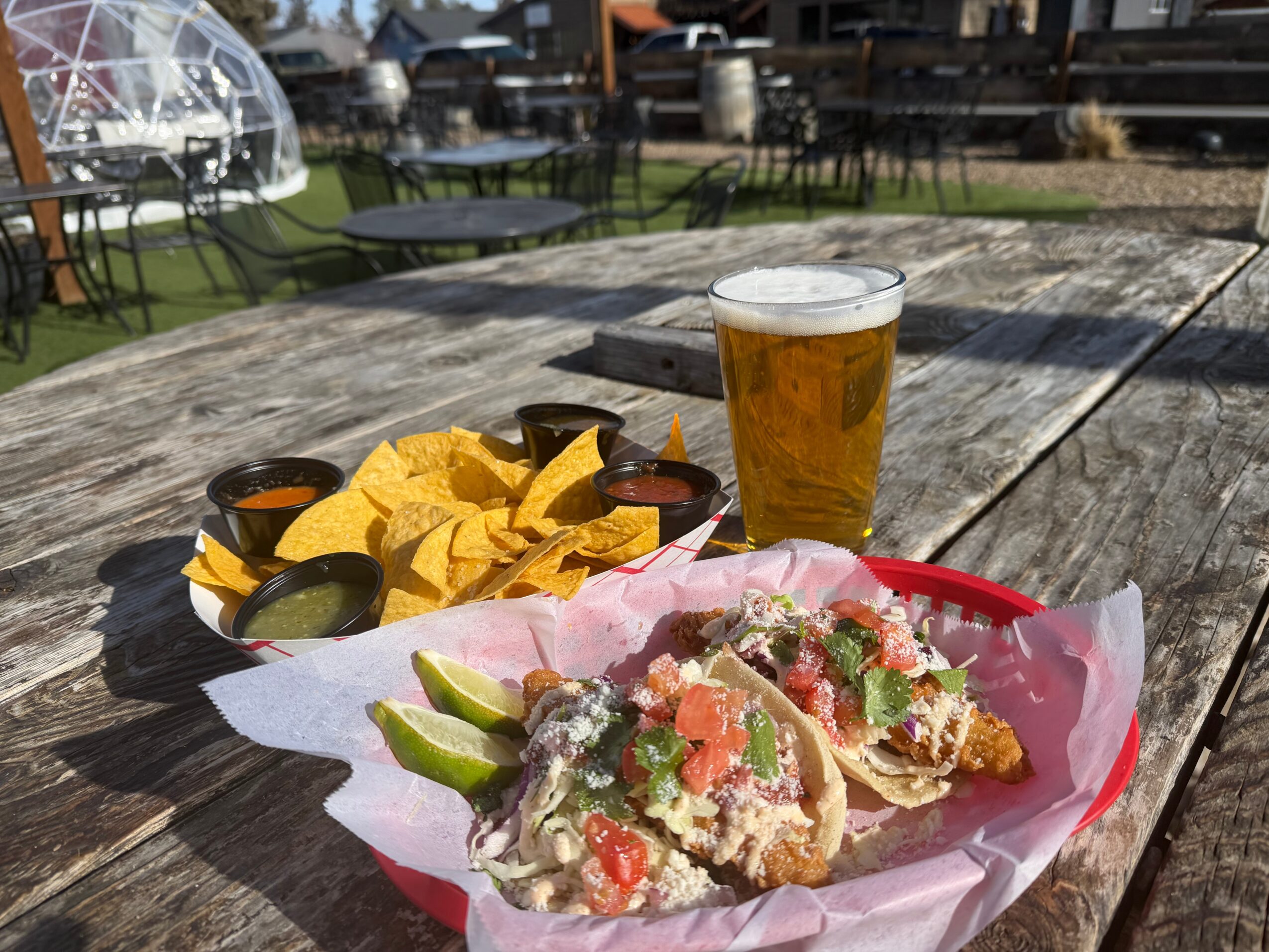 A plate of tacos with lime wedges, a glass of beer, and small sauce containers on a wooden table outdoors.