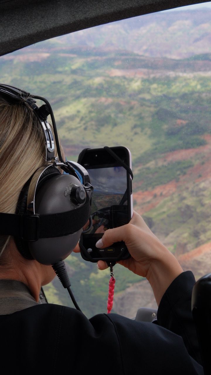 Person wearing headphones and sunglasses, holding a phone with a red cord, looking out of an aircraft window at landscape.