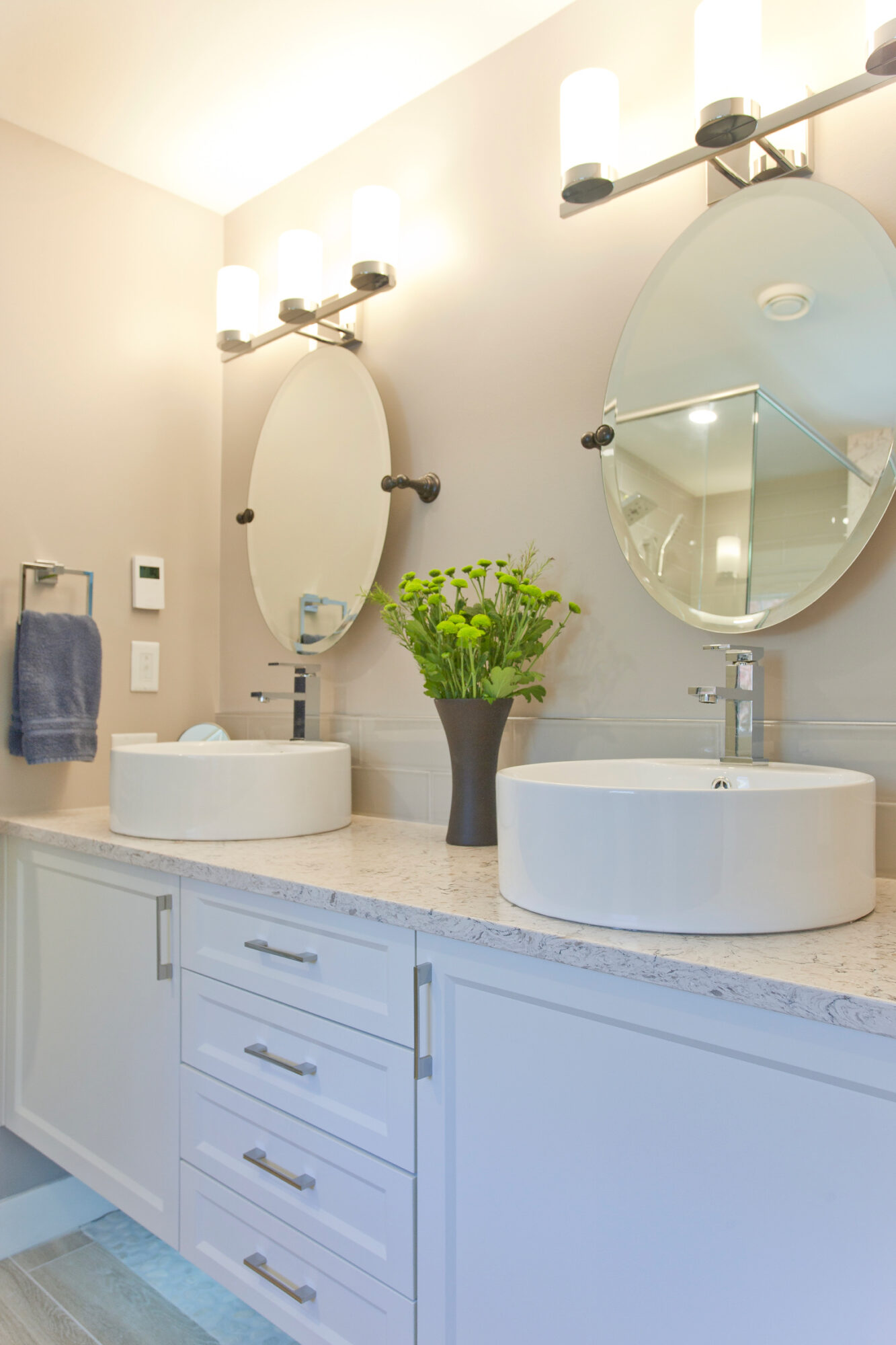 Bathroom with two oval mirrors above sinks, a potted plant, and white cabinetry, illuminated by wall-mounted lights.
