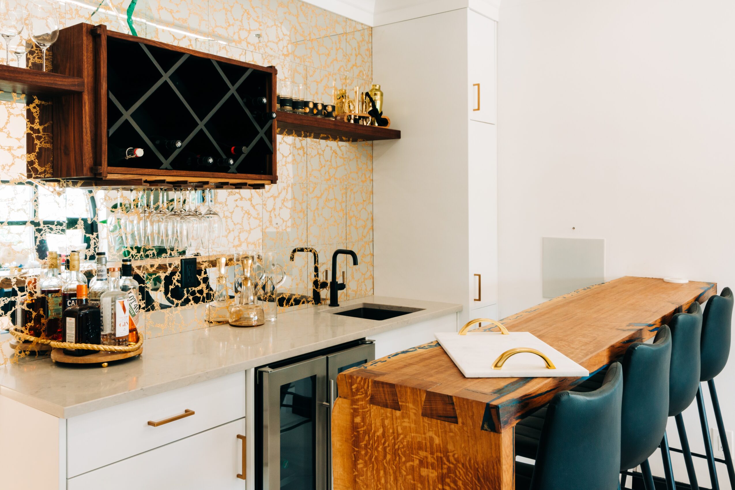 Kitchen with a wooden shelf, wine rack, sink, and bar counter with chairs, open book on counter, and white wall.