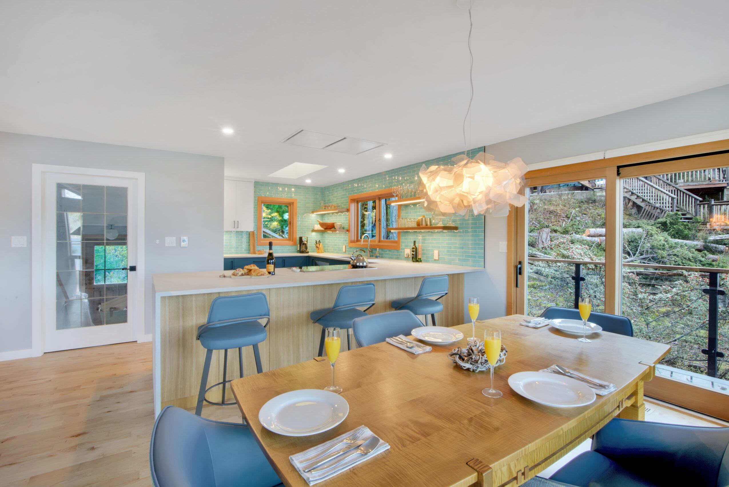 Open kitchen and dining area with large window, wooden table set for four, blue chairs, and a kitchen island with barstools.