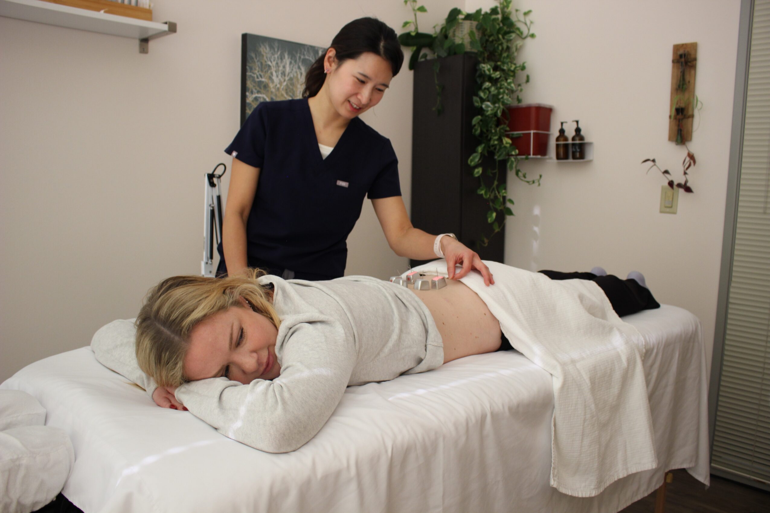 A woman receives a massage from a therapist in a treatment room with plants and shelves, lying face down on a massage table.