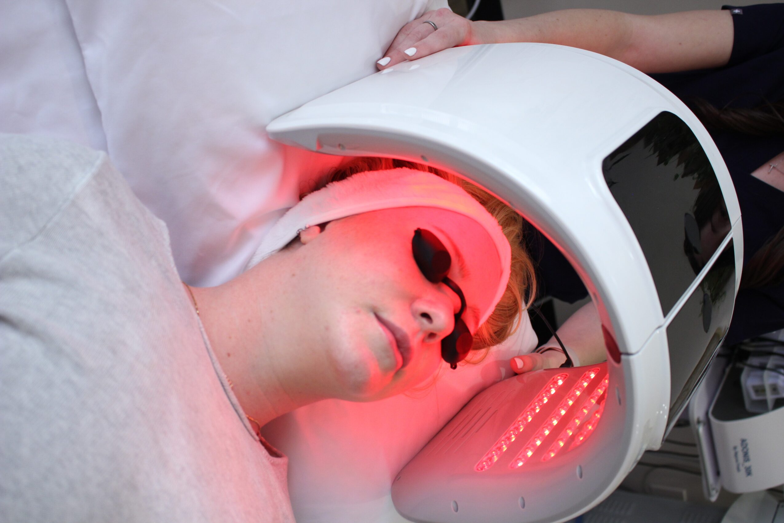 Person receiving a facial treatment with red light therapy, lying down with protective goggles, in a clinical setting.