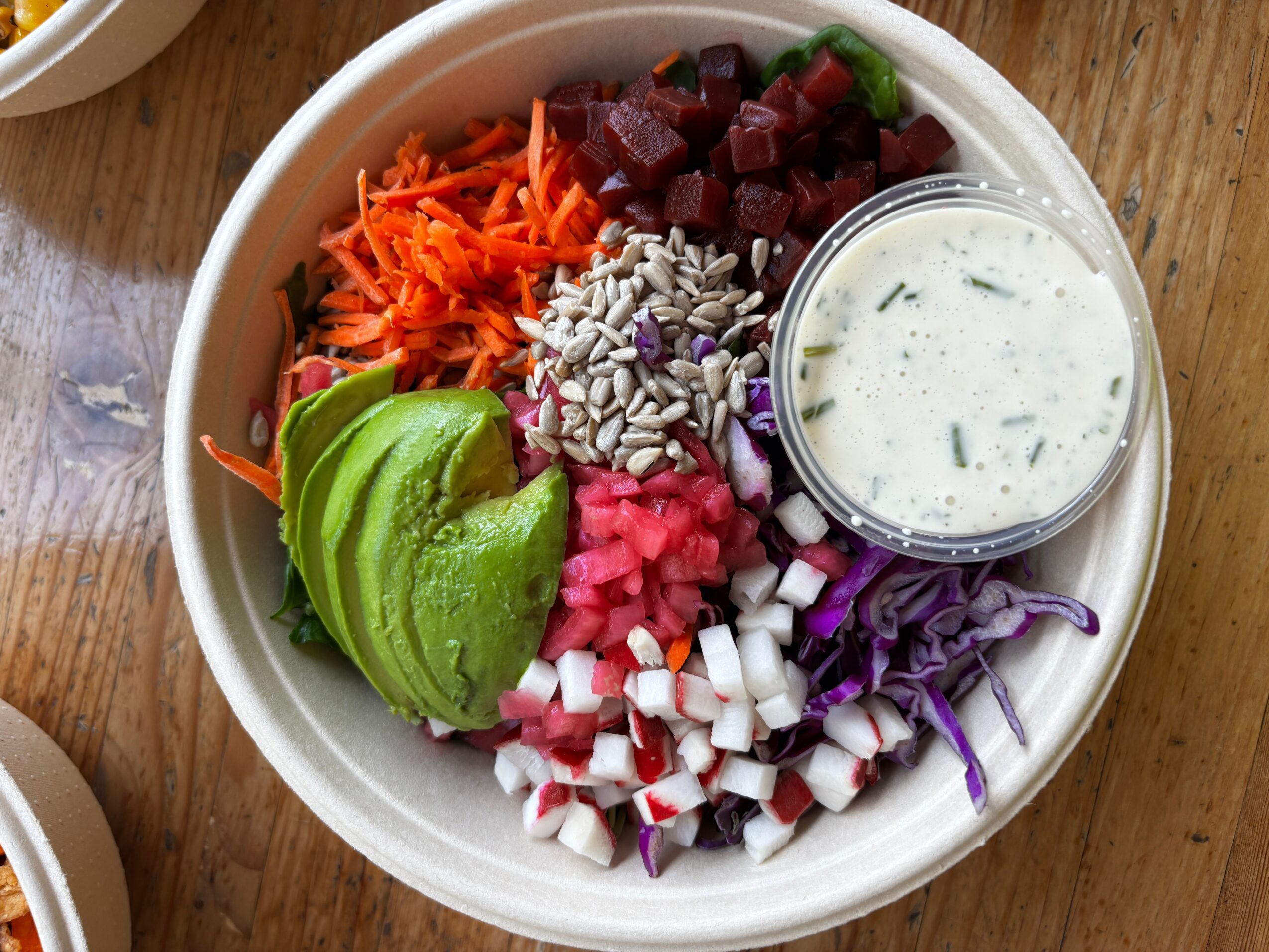 Bowl with chopped vegetables, avocado slices, and a small container of creamy dressing on a wooden surface.